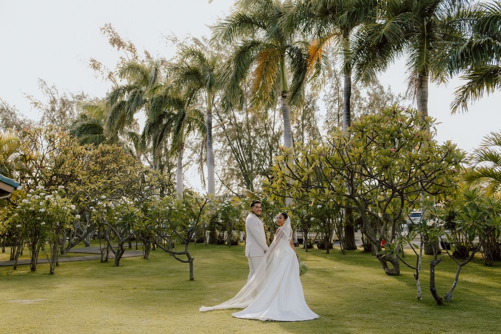 Two brides in white wedding dresses stand together on a lush lawn surrounded by tropical palm trees and greenery.