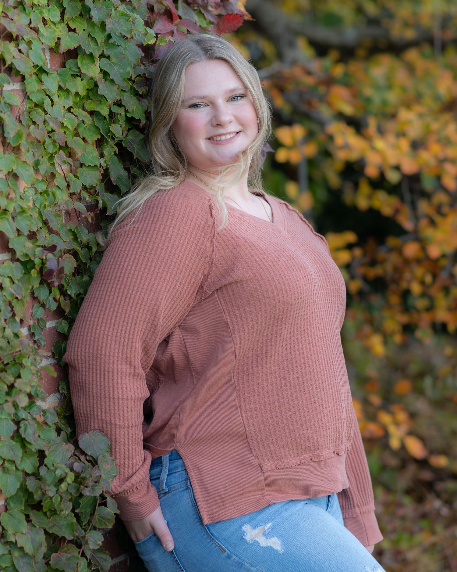 Casual autumn portrait of person in pink sweater leaning against vine-covered wall.
