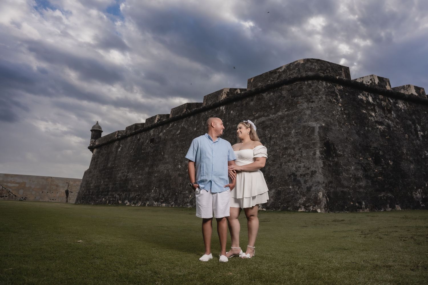 A couple stands together on the green lawn in front of the historic stone walls of Castillo San Felipe del Morro fortress.