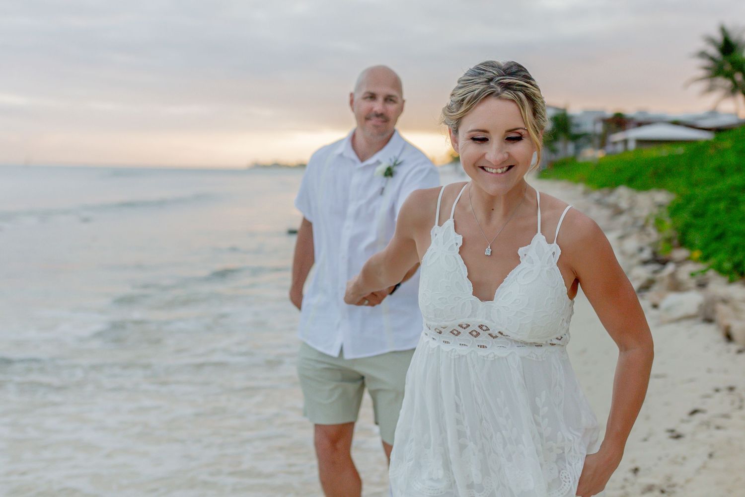 A couple in white outfits stands on a beach at sunset with palm trees and waves in the background.
