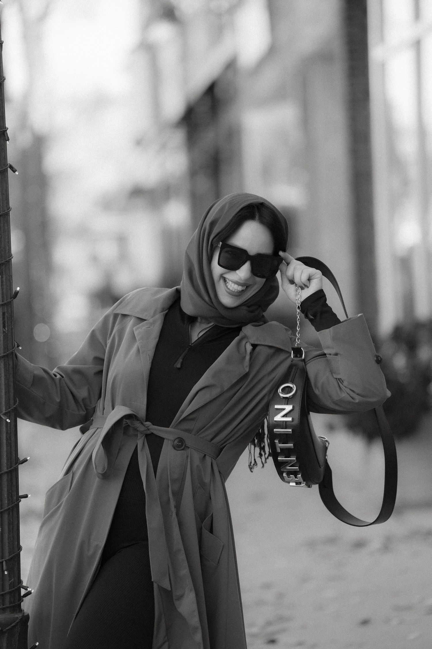 A person in a trench coat and headscarf poses stylishly on an urban street.
