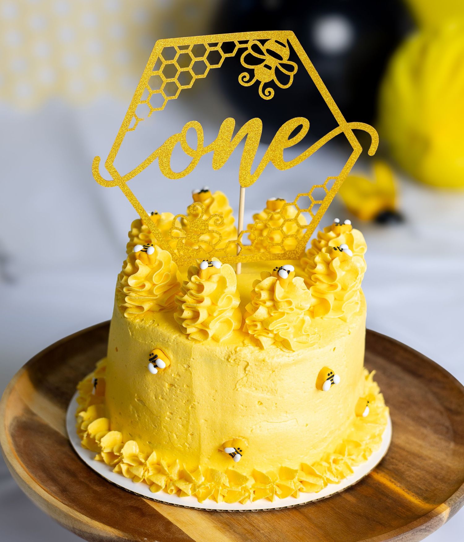 Yellow honeycomb-themed cake with gold 'one' topper and decorative piping on wooden plate.