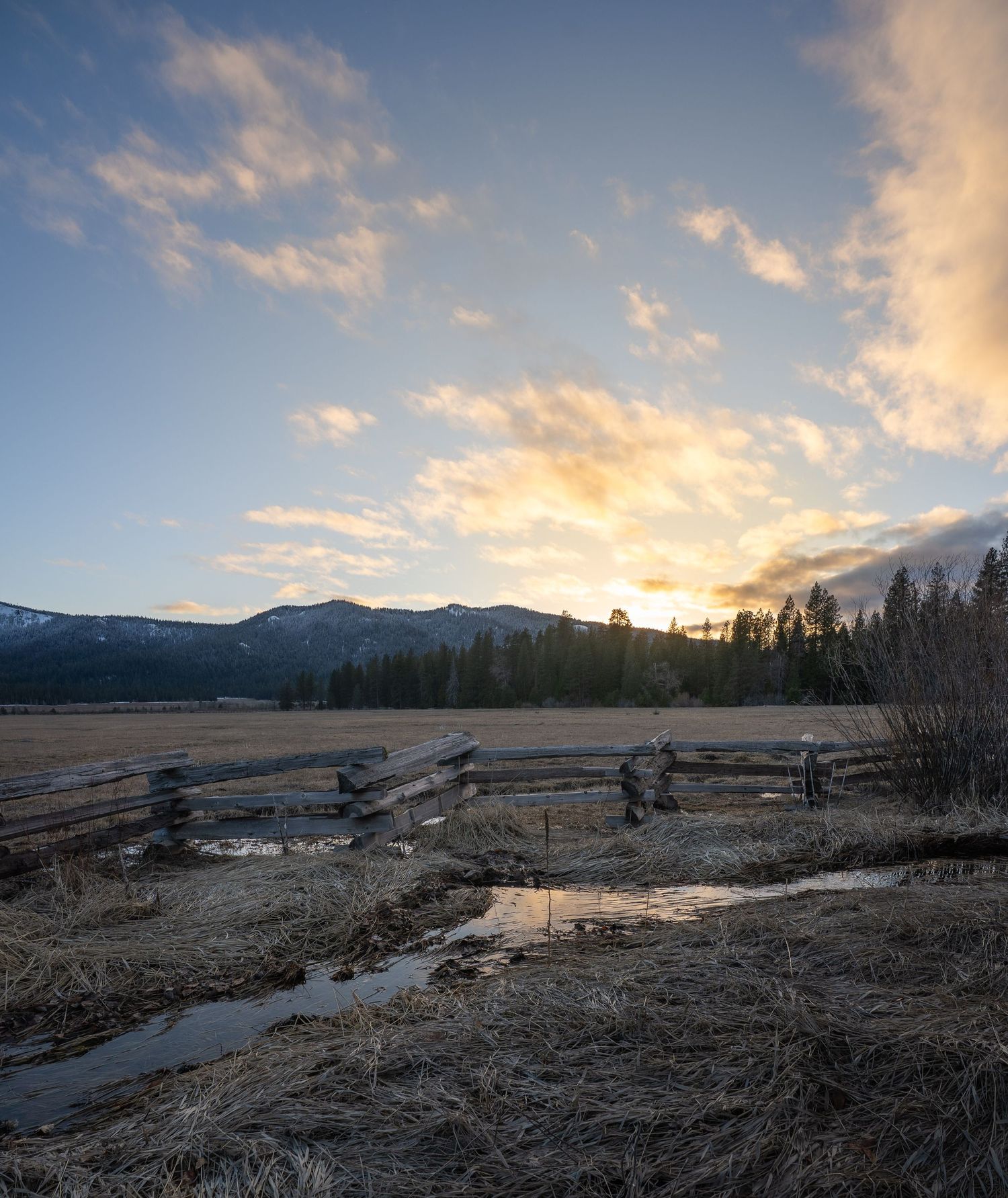 Mountain landscape at sunset with rustic wooden fence and melting snow reflecting golden light in early spring.