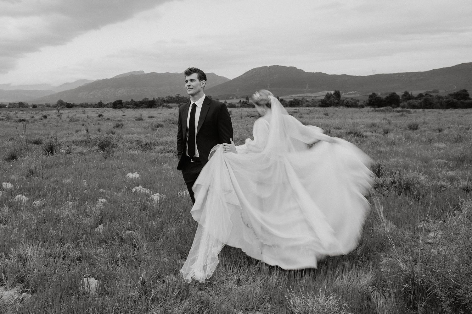 Black and white wedding photo showing a couple in formal attire standing in a dramatic mountain meadow landscape.