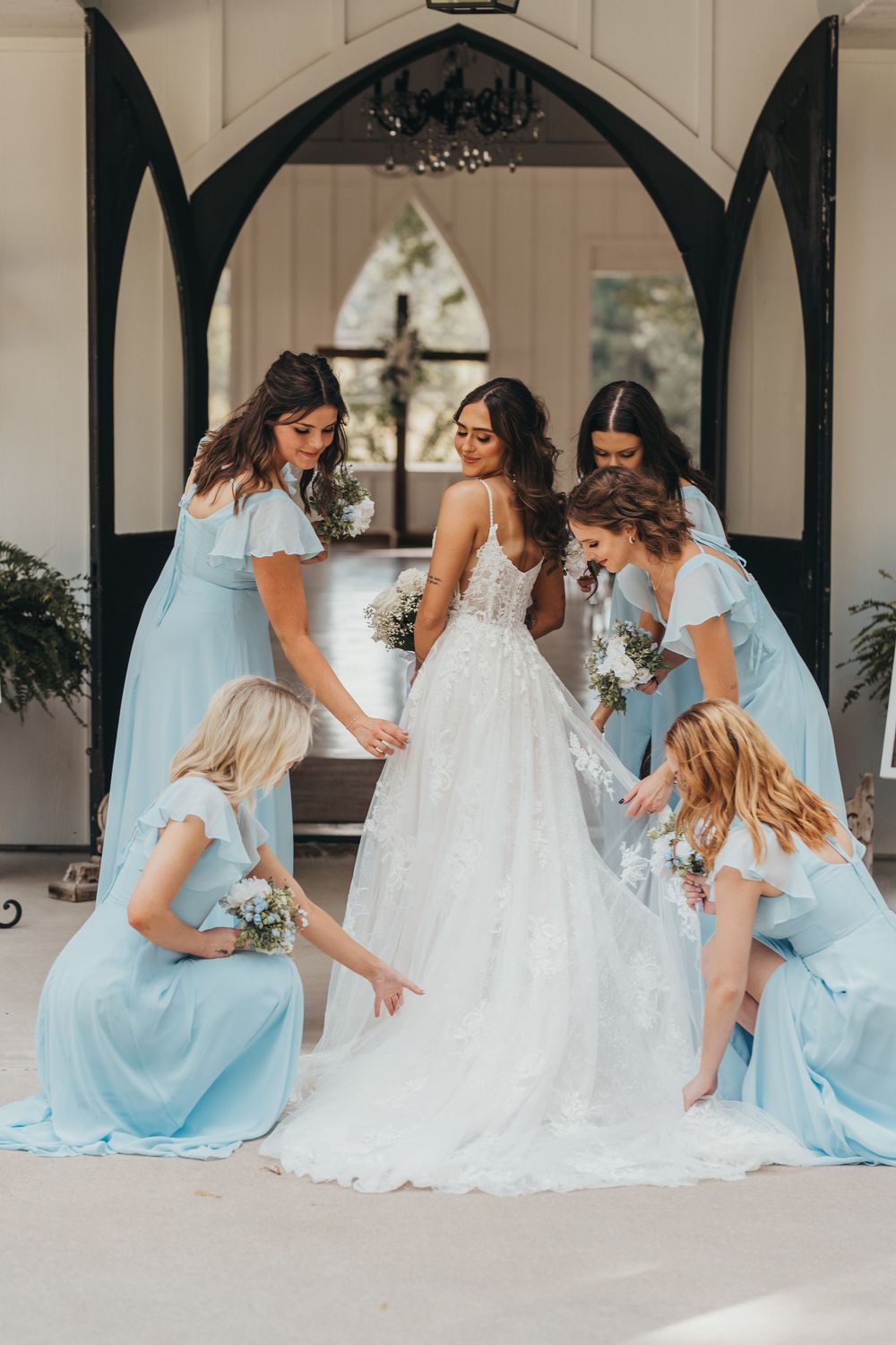Bridesmaids in light blue dresses help adjust the bride's white gown while sitting on a church porch.