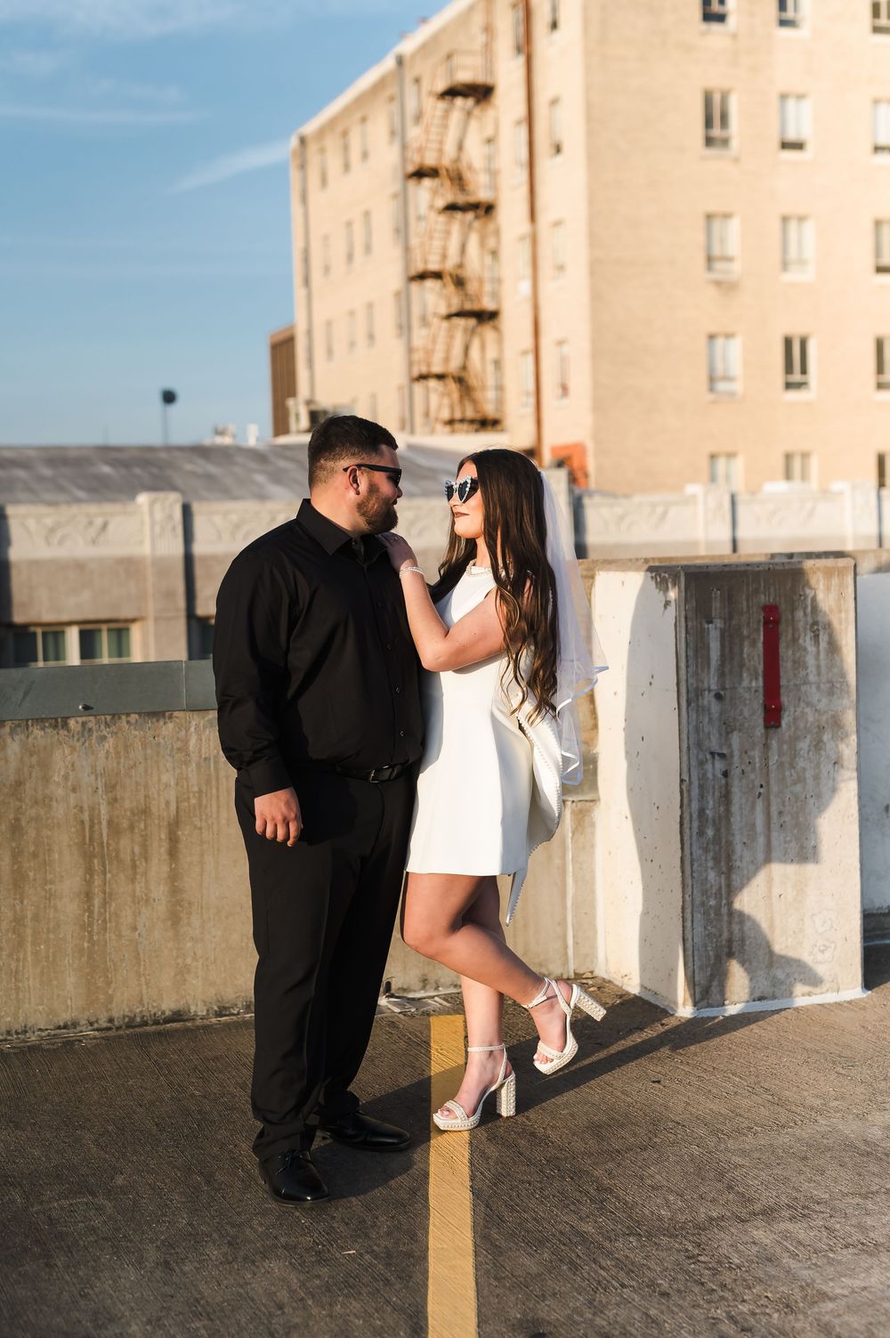 A couple in contrasting black and white outfits stand together on a rooftop parking structure with an apartment building behind them.