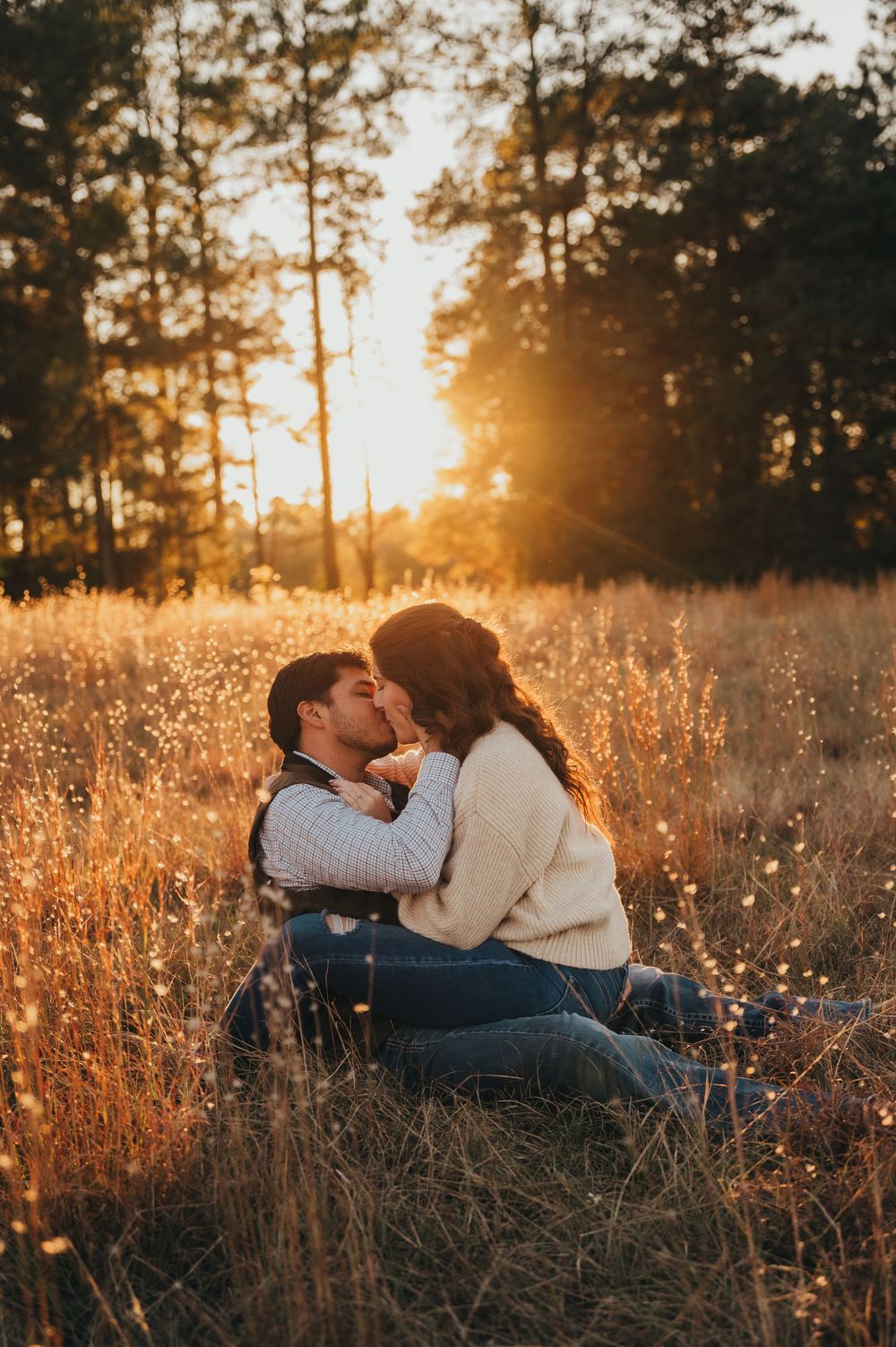 Romantic couple lying in golden sunlit meadow with tall grass and pine trees at sunset.
