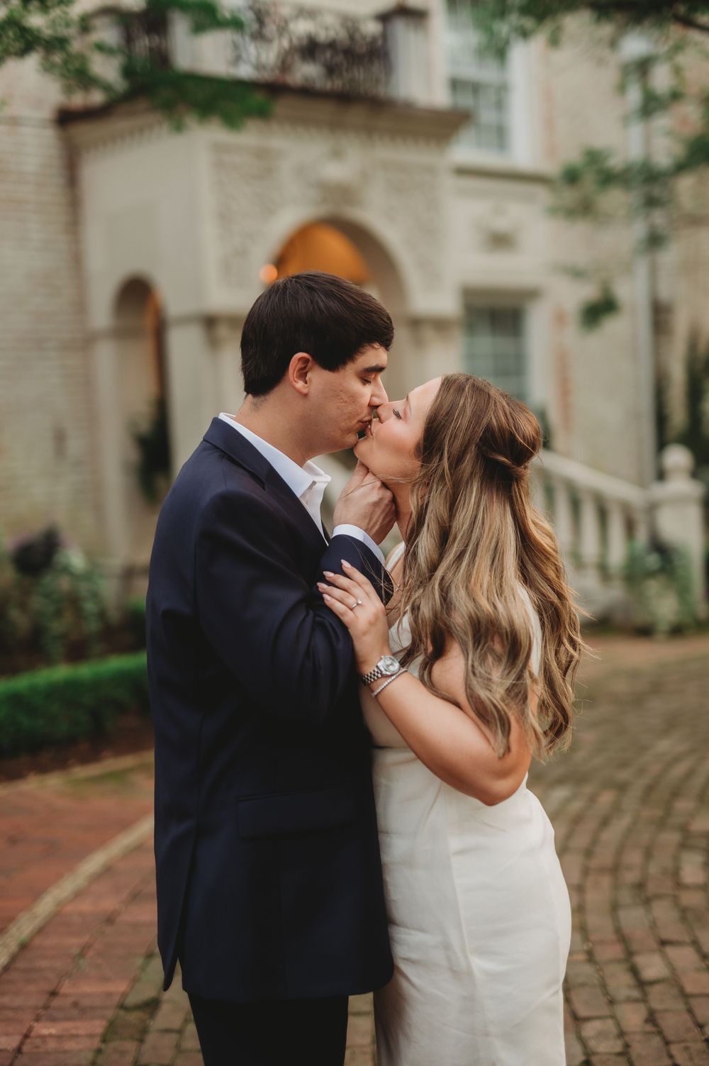 A couple shares a romantic kiss outdoors, the groom in a navy suit and bride in a white dress, with a elegant building backdrop.