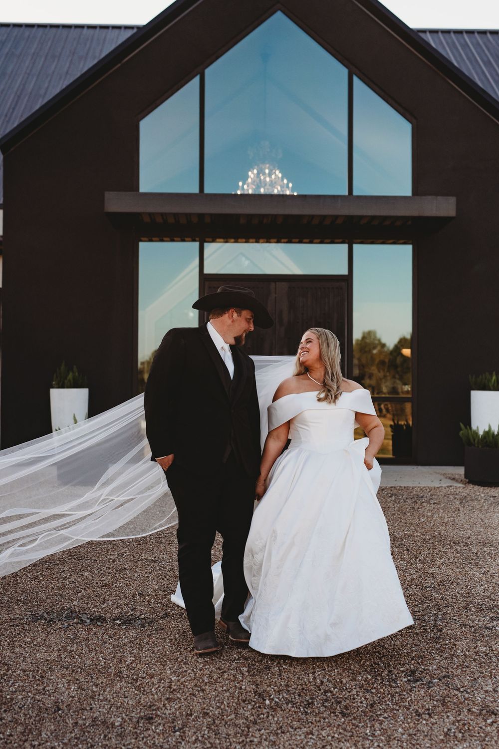 A wedding couple stands outside a modern black chapel with the bride's veil flowing dramatically in the wind.