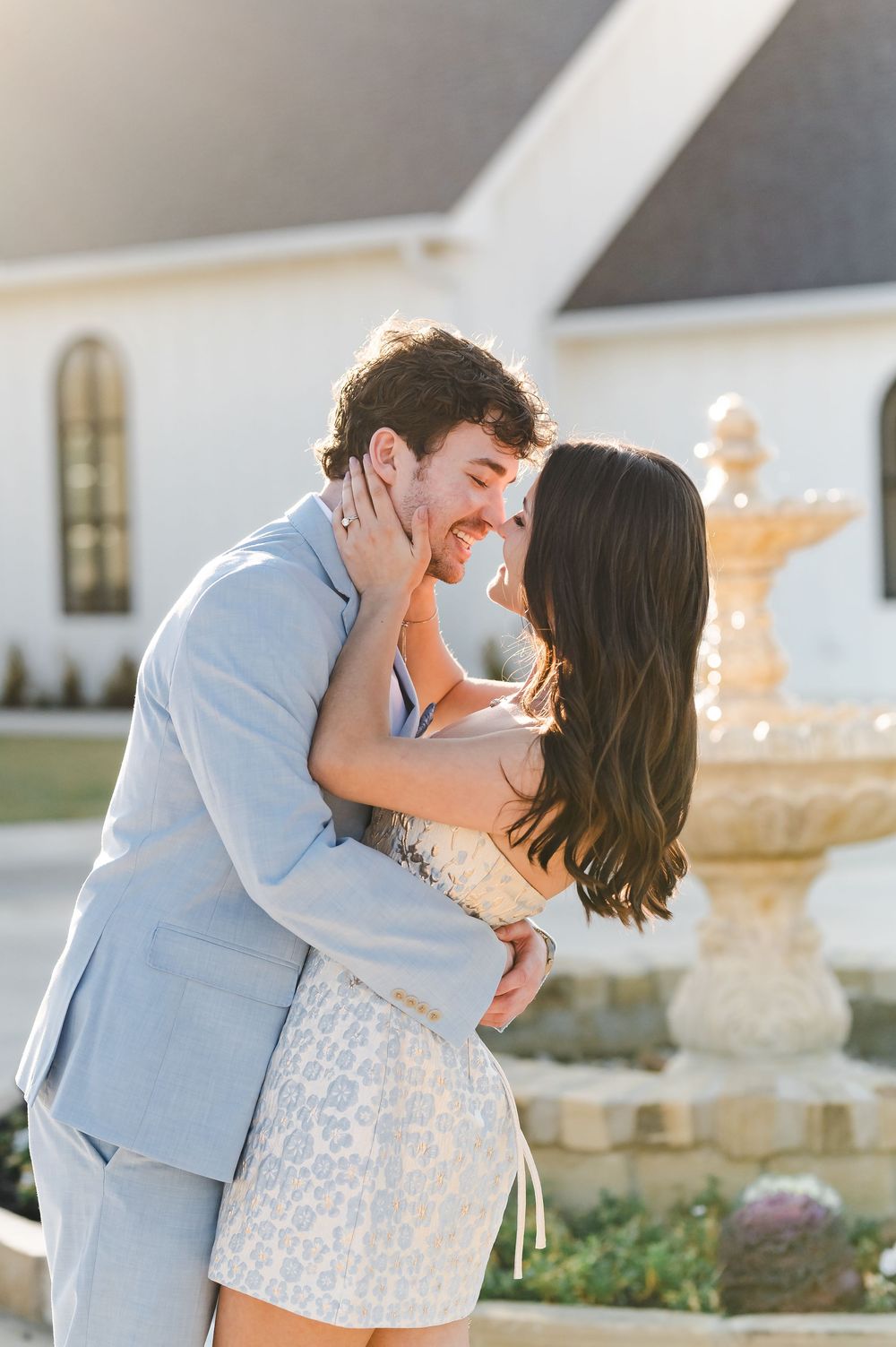 A romantic couple shares tender kisses in a garden setting while wearing light blue and white outfits at sunset.