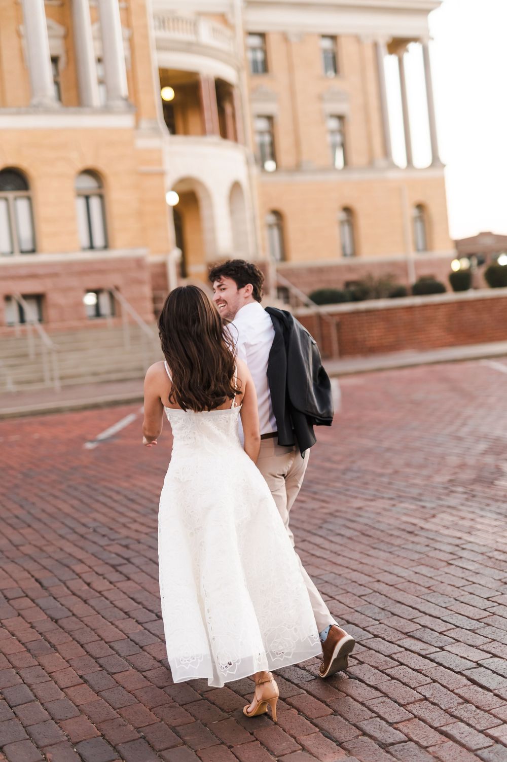 A couple in wedding attire dance and twirl on a brick courtyard outside a historic building.
