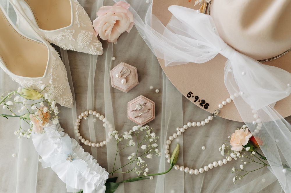 Elegant wedding flat lay with bridal heels, pearl bracelet, ring box, veil, and floral accents on soft fabric.