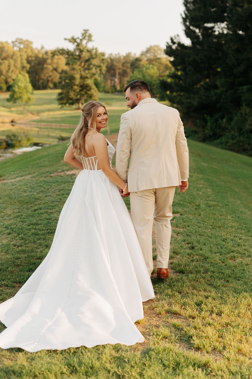Bride in white gown and groom in tan suit walk hand-in-hand across lush green lawn at golden hour.