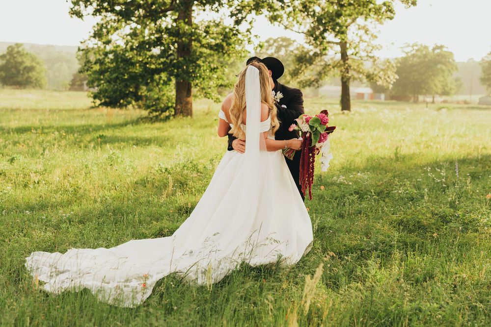Bride in white gown with train and groom in black hat walking through lush green meadow on wedding day.