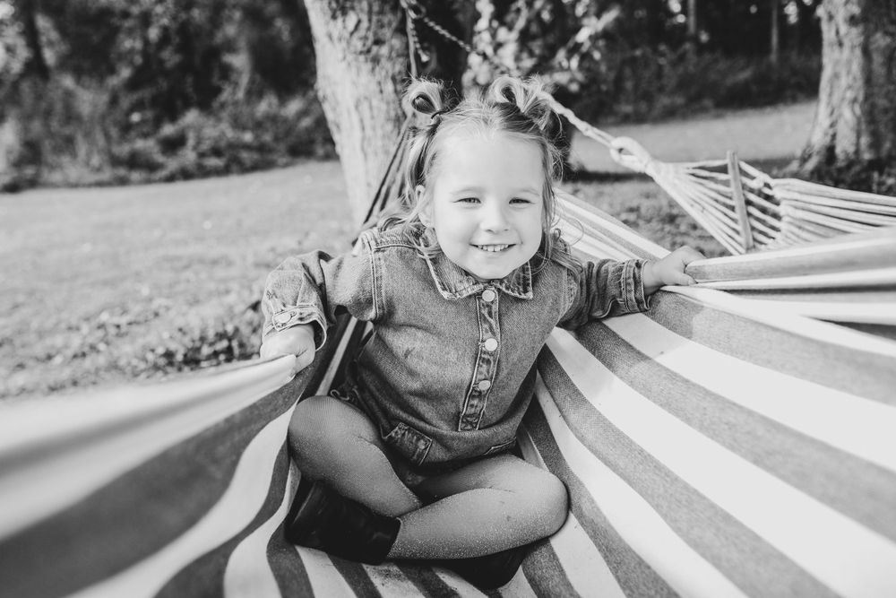 Black and white portrait of a smiling child sitting on striped hammock in backyard.