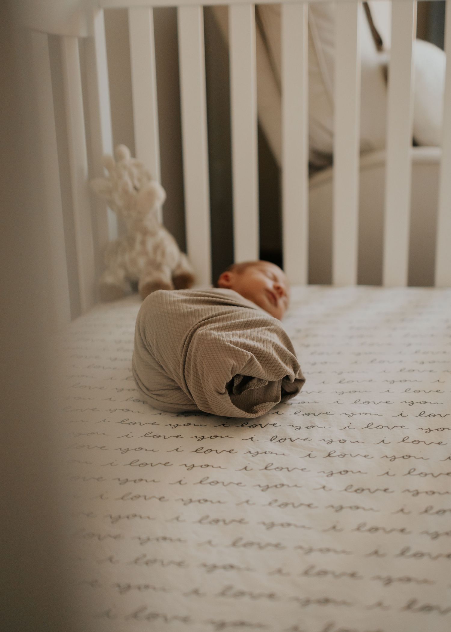 A sleeping infant in gray swaddle rests on bedding with repeating 'I love you' text pattern in a white crib.