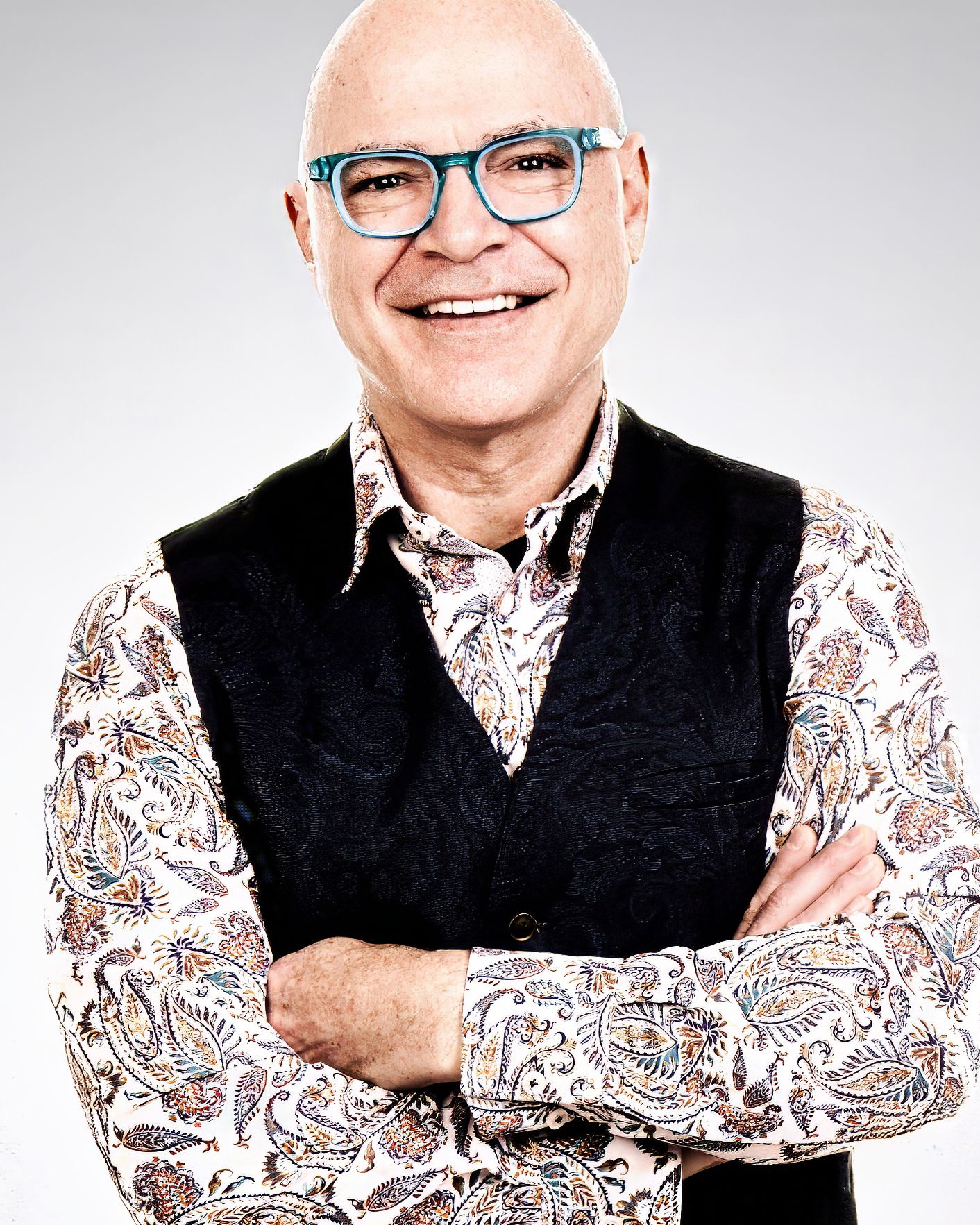 Professional portrait of someone wearing glasses, a floral print shirt and black vest, standing with arms crossed and smiling.