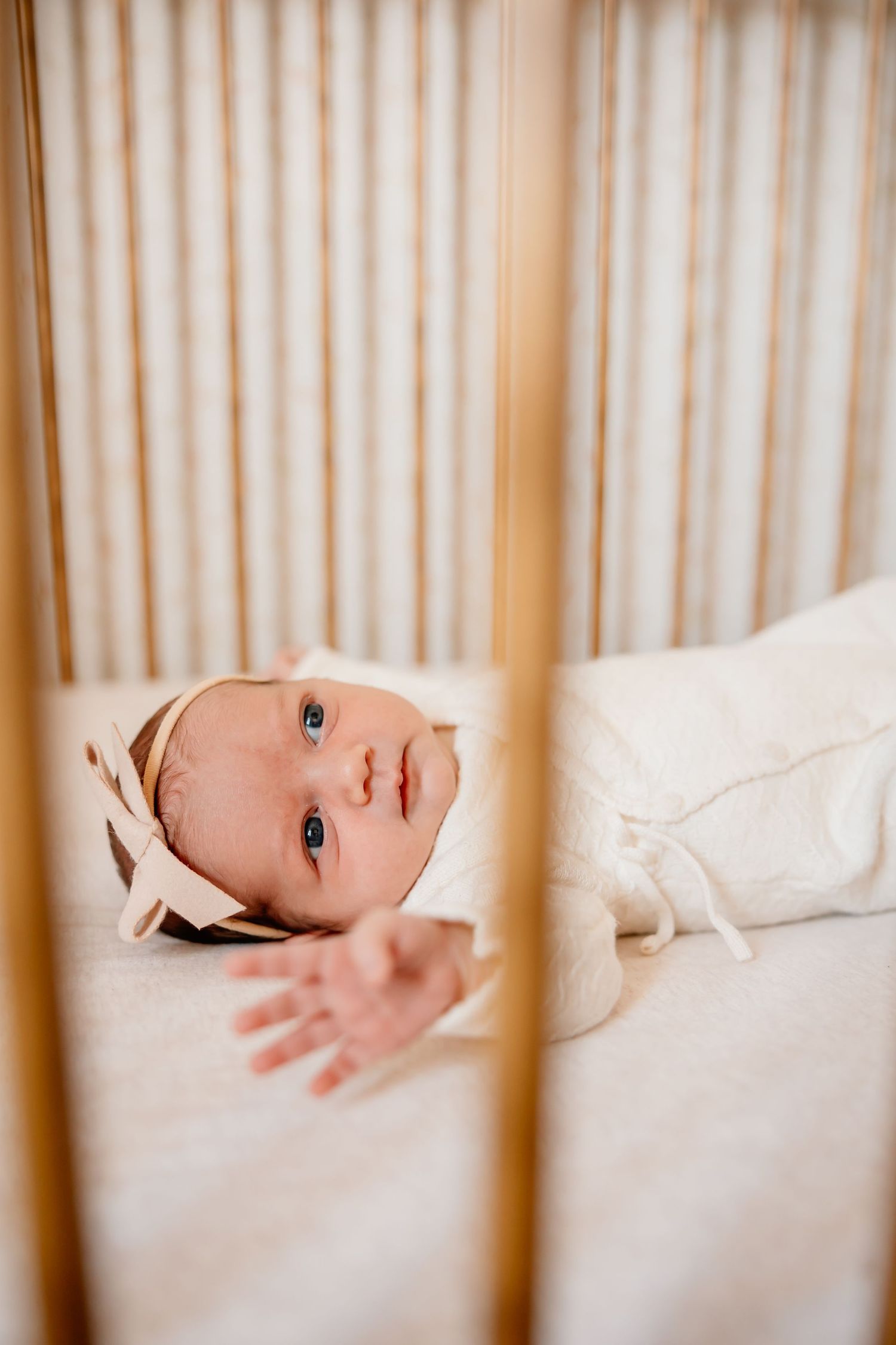 Baby wearing white bonnet lying in wooden crib looking through slats.