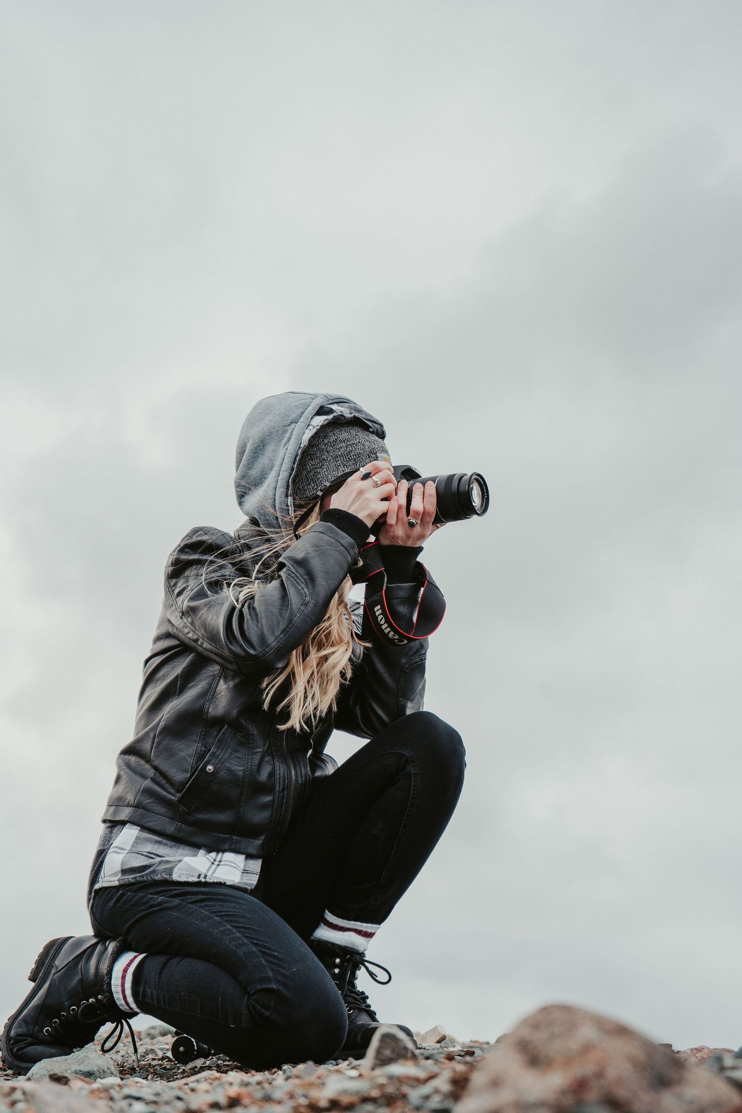 A photographer in a gray hoodie kneels on rocky ground while taking photos with a telephoto lens against an overcast sky.
