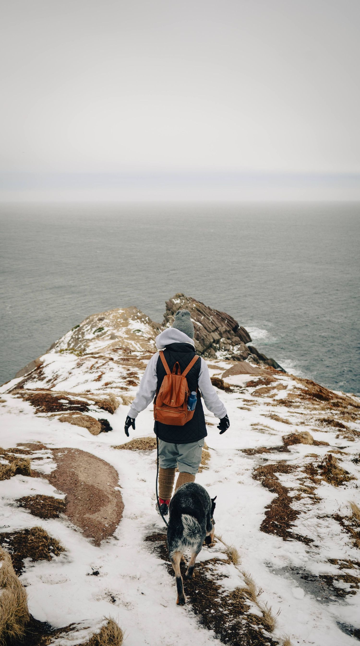 Hiker with orange backpack walks along snowy coastal trail overlooking misty ocean horizon on winter day.