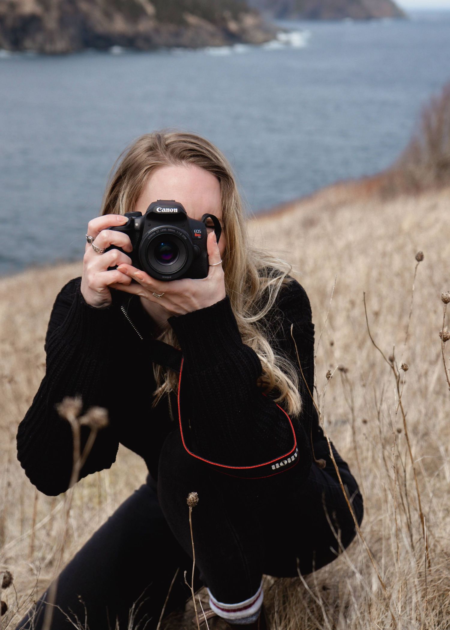 Photographer with black sweater takes pictures on coastal cliffs with dried grass and ocean waves in background.