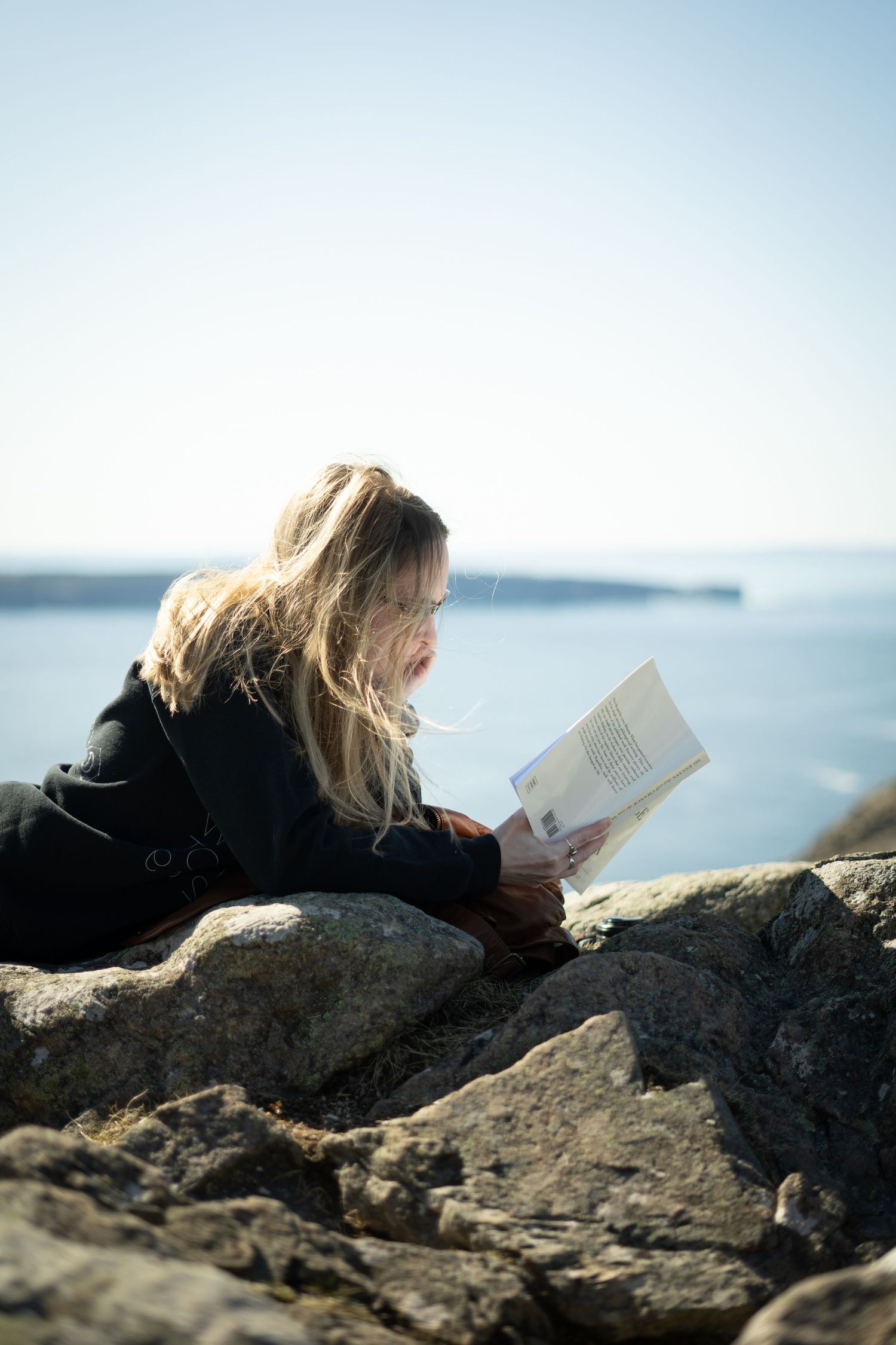 Reader enjoys a peaceful moment on coastal rocks with a book while overlooking calm ocean waters on a bright day.