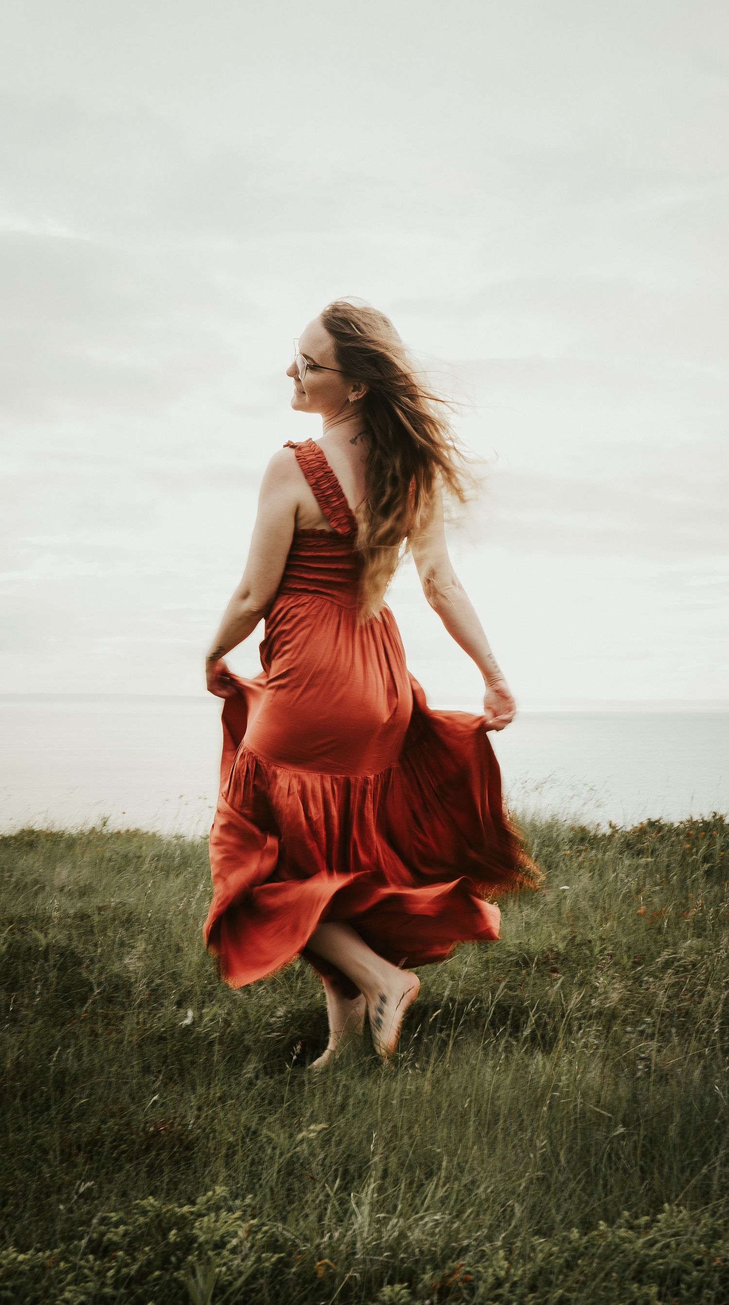 A woman in a flowing red dress dances barefoot through a windswept meadow at dusk.