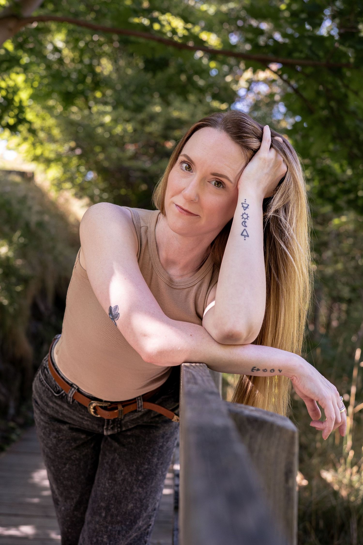 Woman with tattoos leans on wooden railing in sunlit garden, posing confidently in beige top and gray jeans.