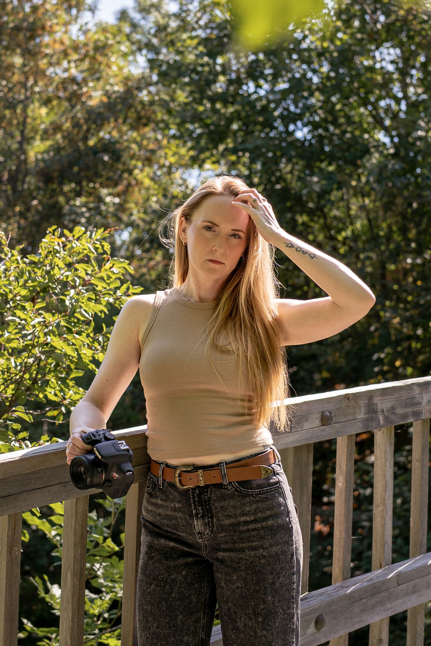 Young woman with long hair holds a camera on a sunny wooden deck surrounded by lush green trees.