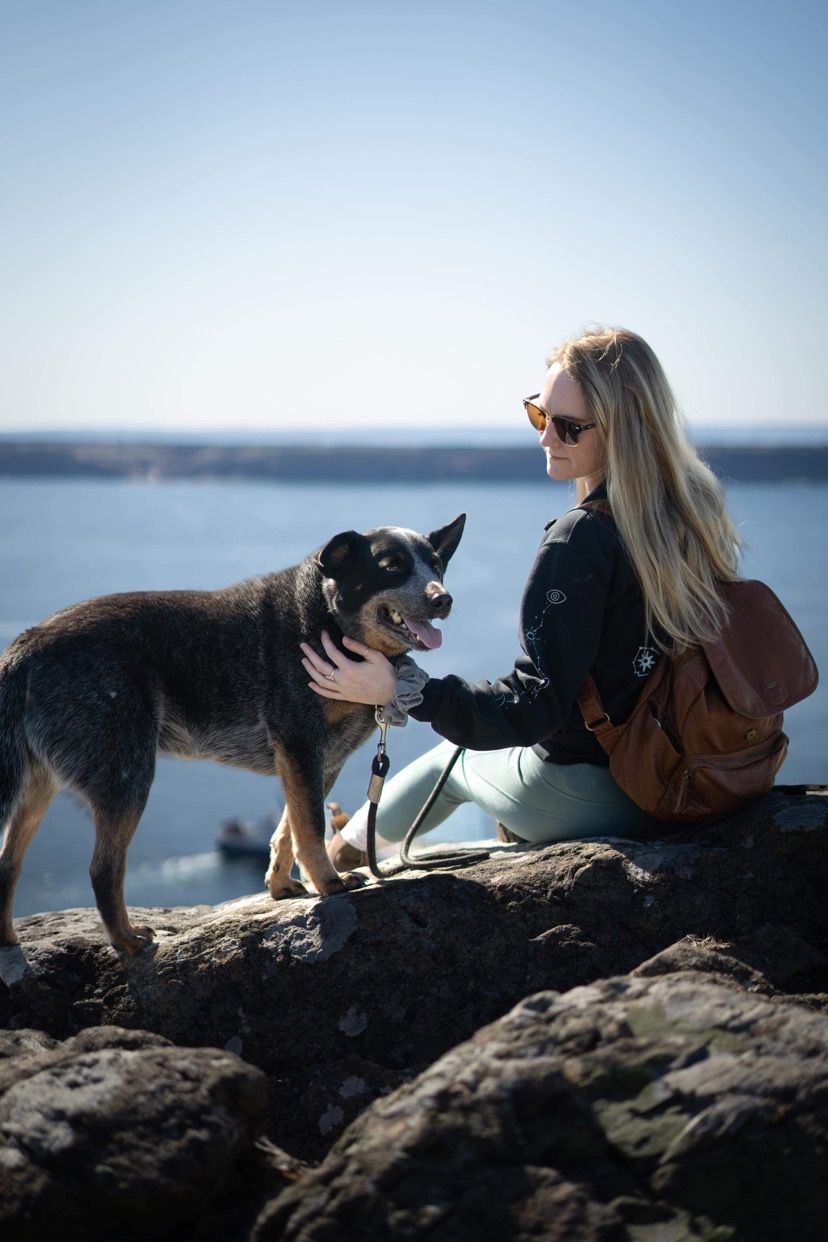 Hiker with long hair pets a black and brown dog while sitting on rocky overlook with scenic water view.