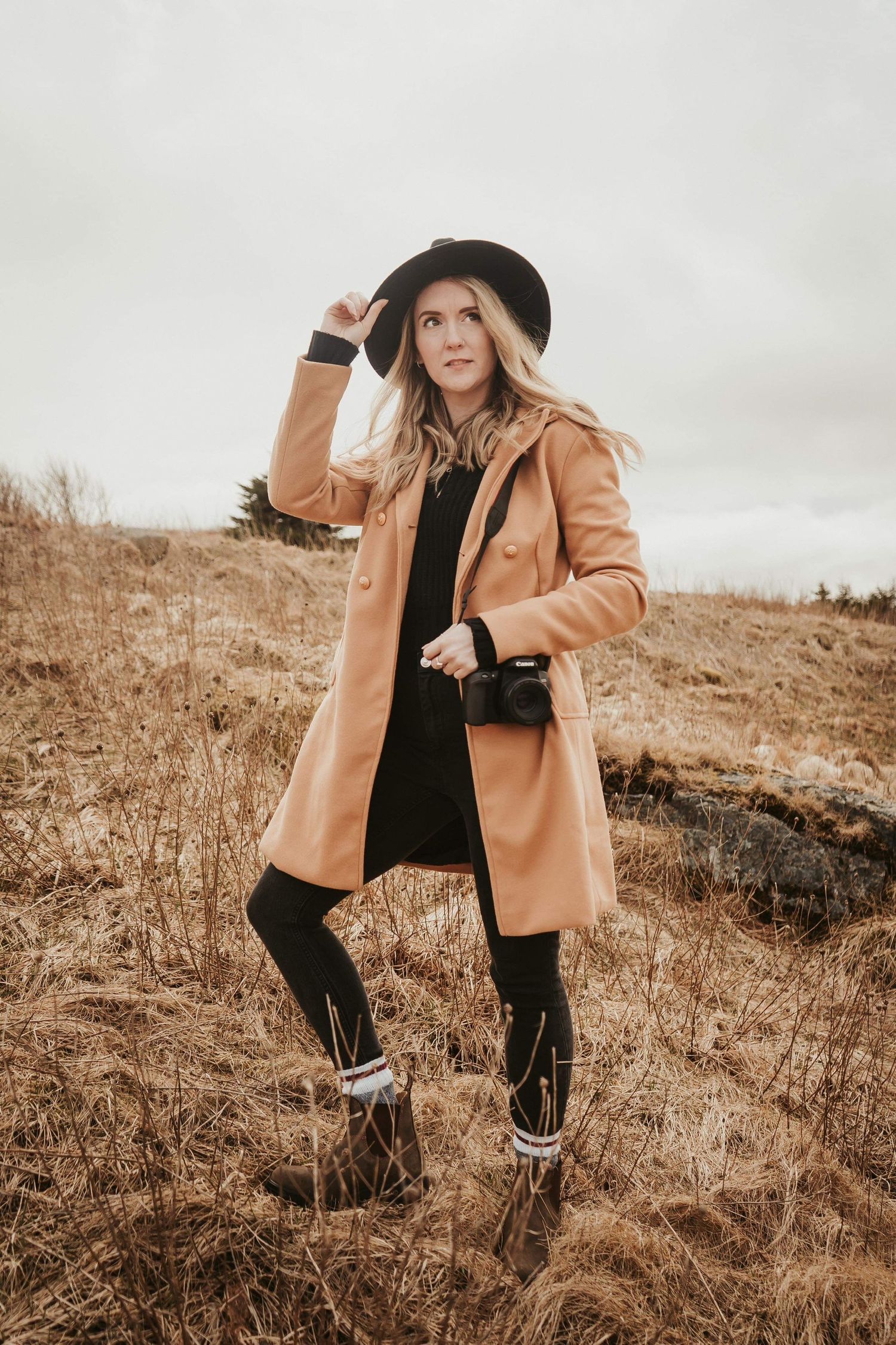 A model in a camel coat, black dress and hat poses on a brown winter hillside in moody natural lighting.