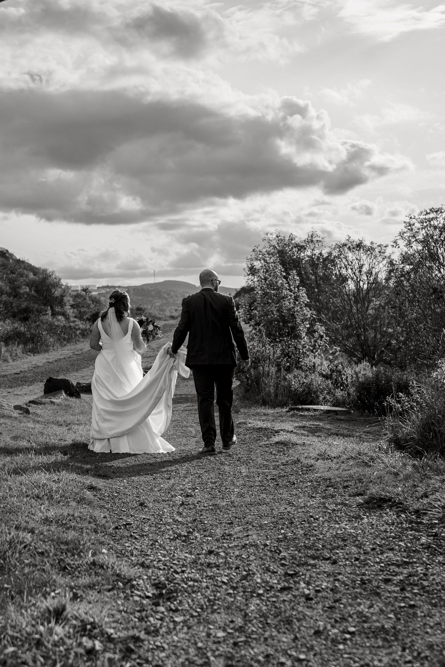 A bride and groom walk hand in hand down a scenic mountain trail under dramatic cloudy skies in black and white.