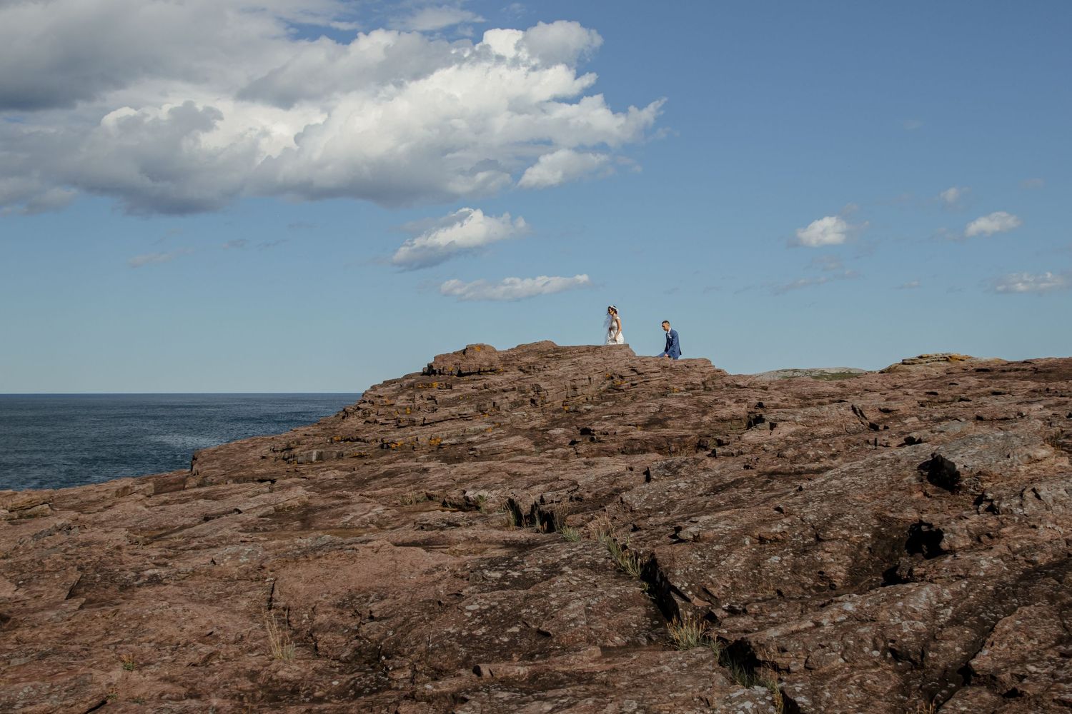 A red rocky coastal shoreline with a lighthouse and figures silhouetted against a blue sky with puffy clouds.