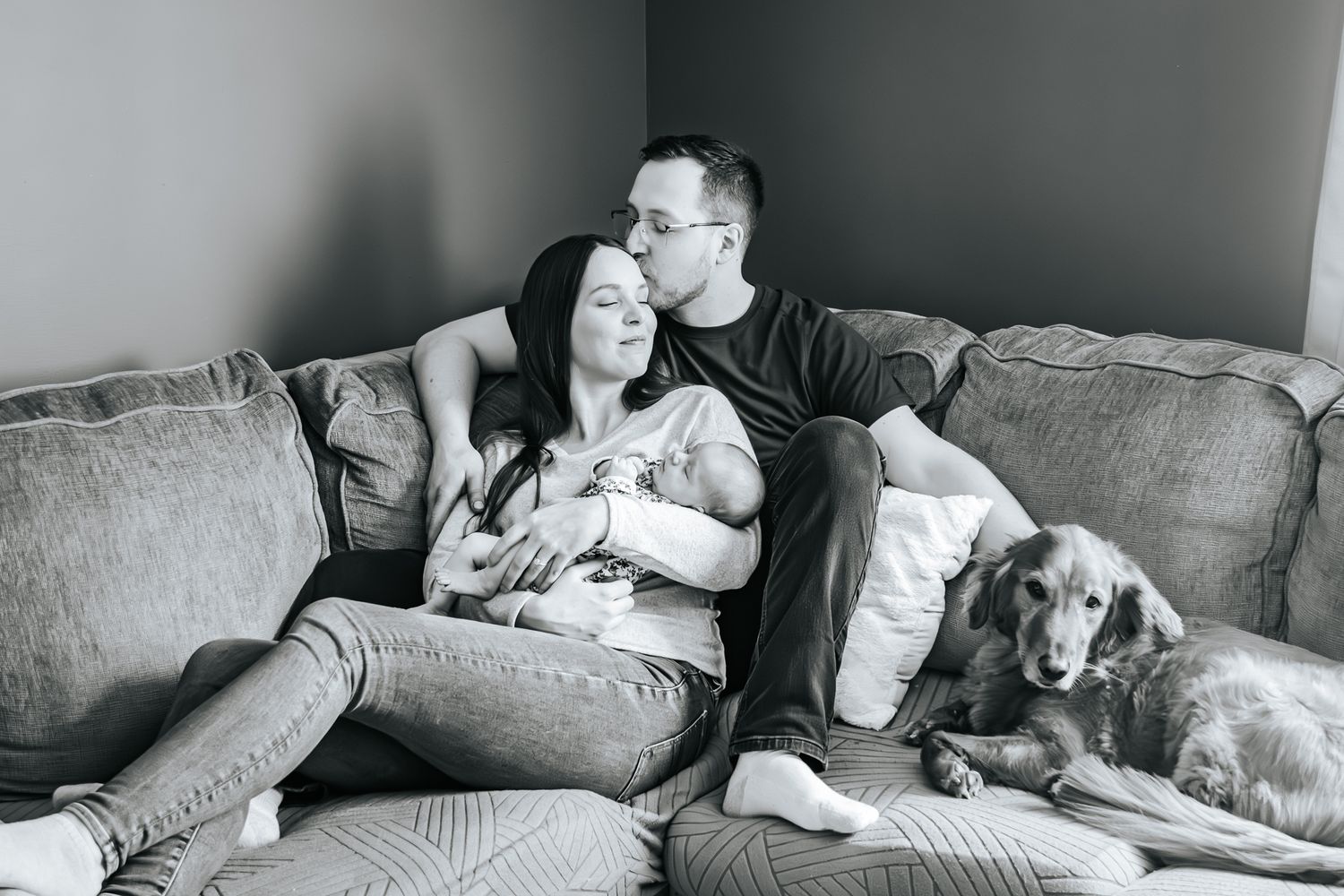 A couple lounges on a cozy gray couch with their two dogs in a candid black and white photograph.