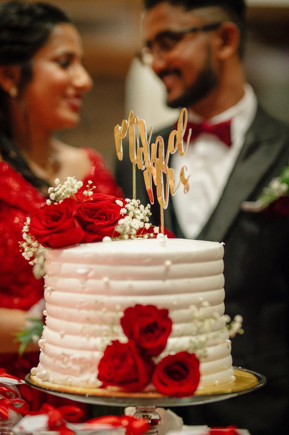 White wedding cake decorated with red roses and gold cake topper at reception.