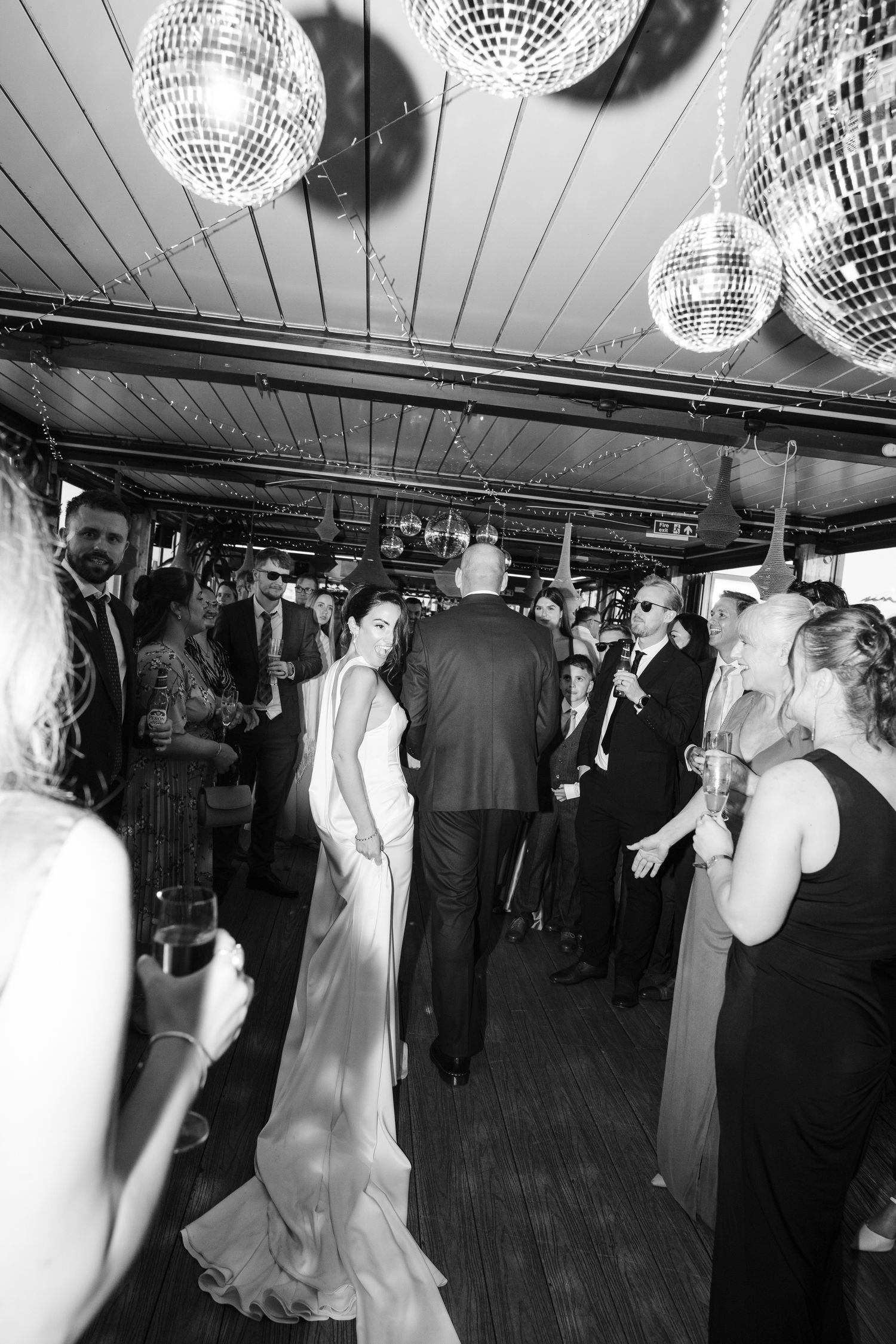A couple in wedding attire dance together in a garden setting at sunset, captured in black and white.