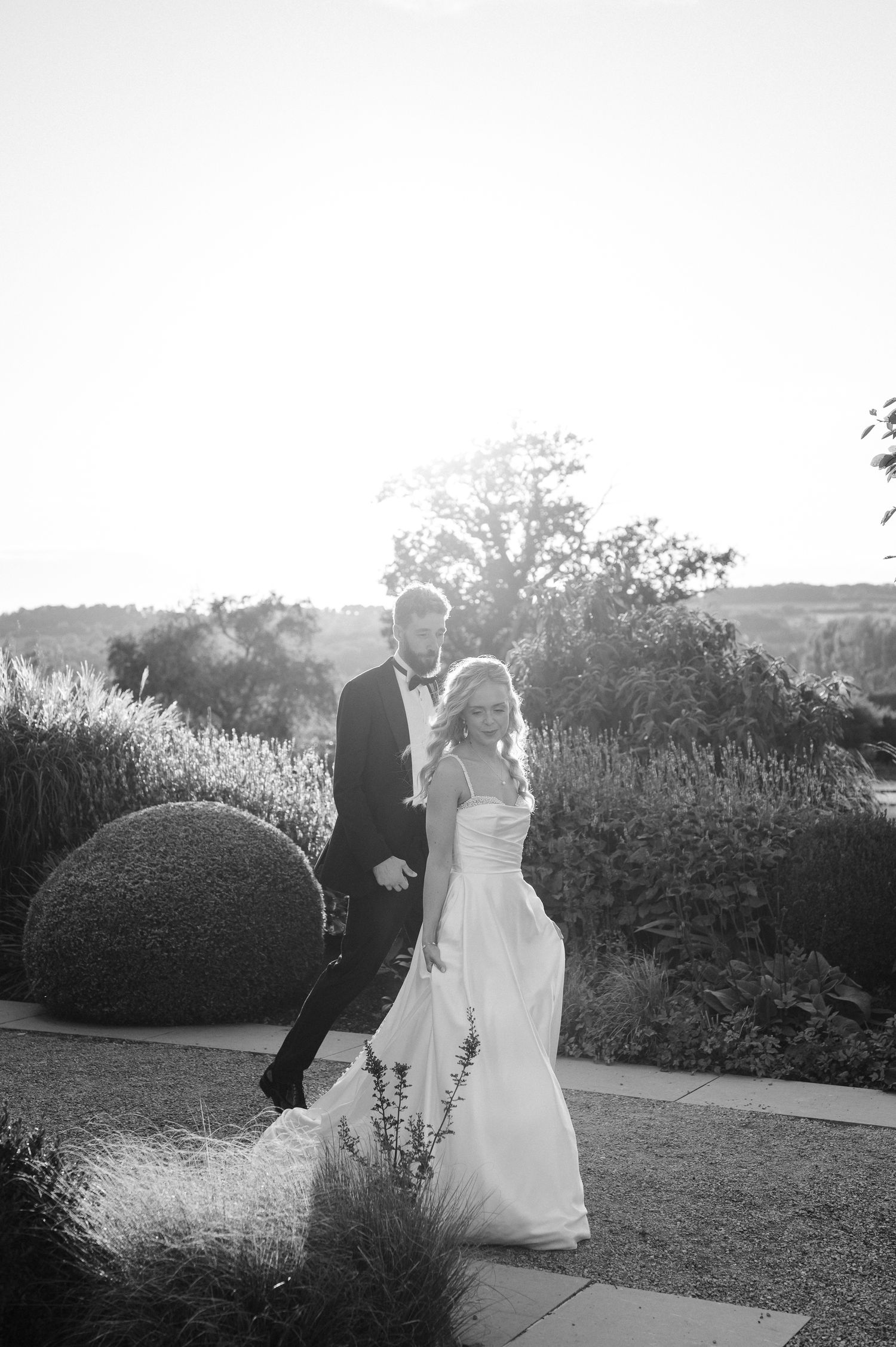 A couple in wedding attire dance together in a garden setting at sunset, captured in black and white.