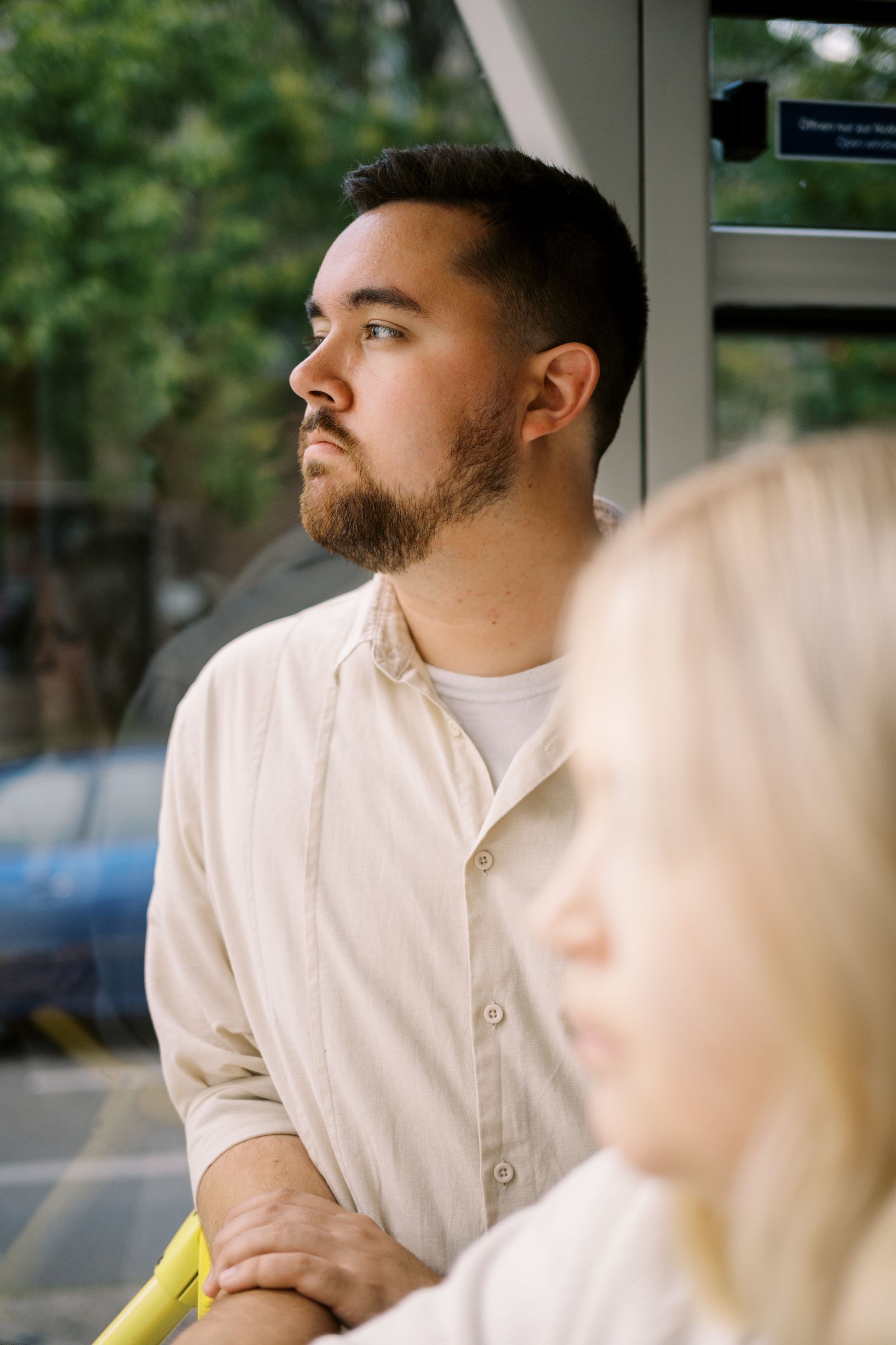 A person with a short beard in a white button-down shirt gazes contemplatively to the side in a bright indoor setting.