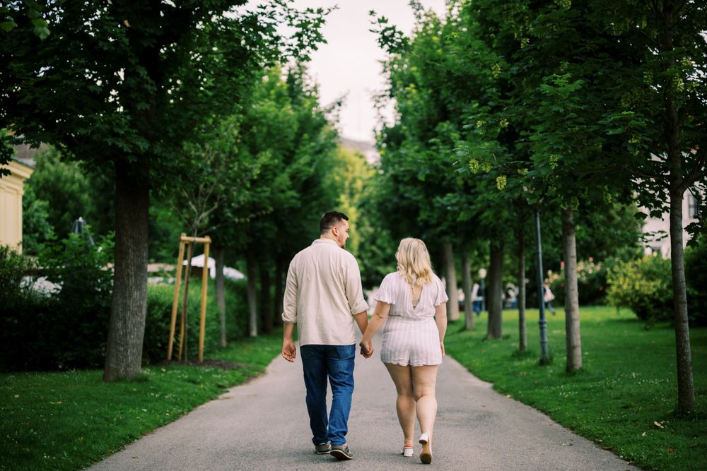 A couple strolls together along a tree-lined path in a lush green park on a summer day.