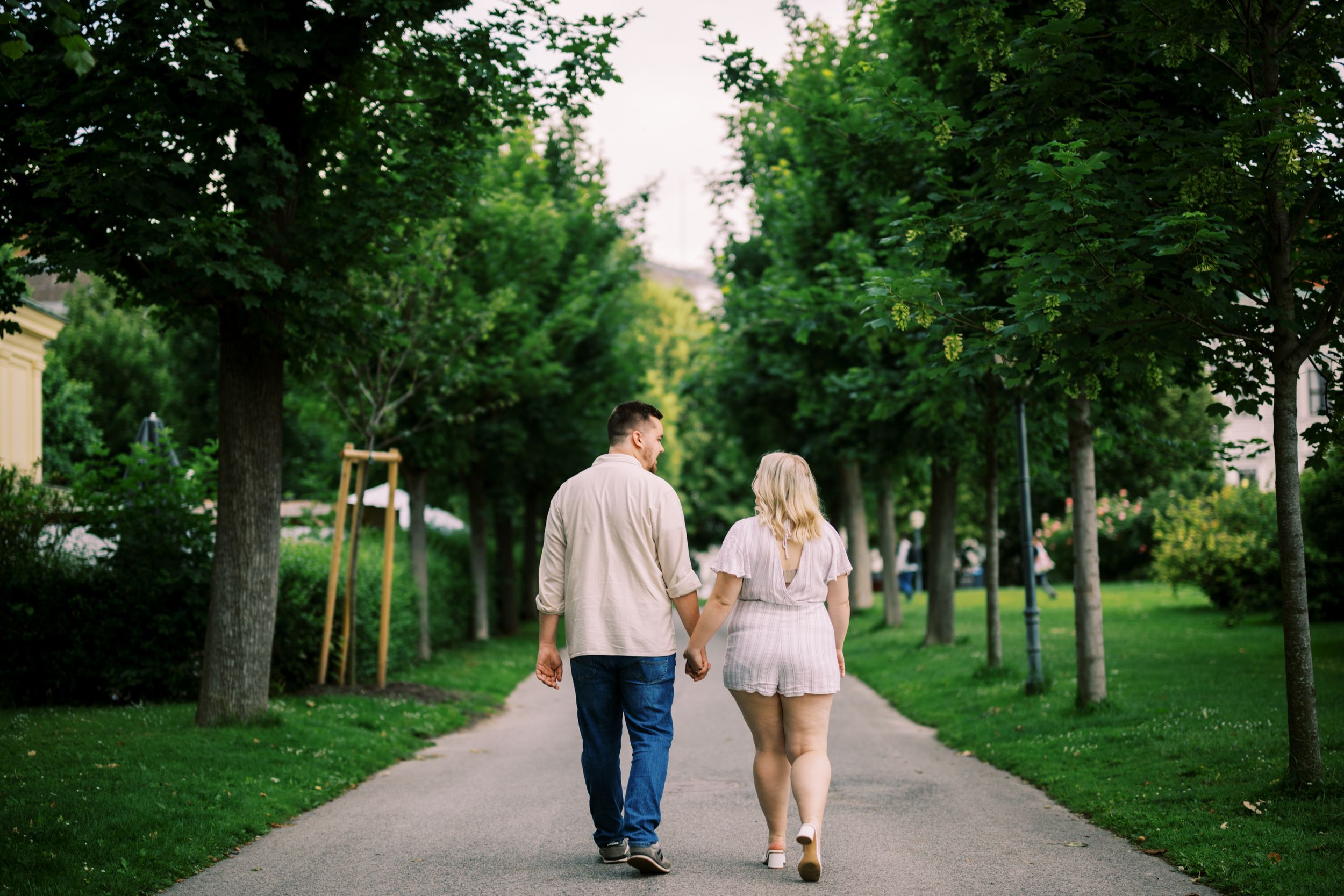 A couple strolls together along a tree-lined path in a lush green park on a summer day.