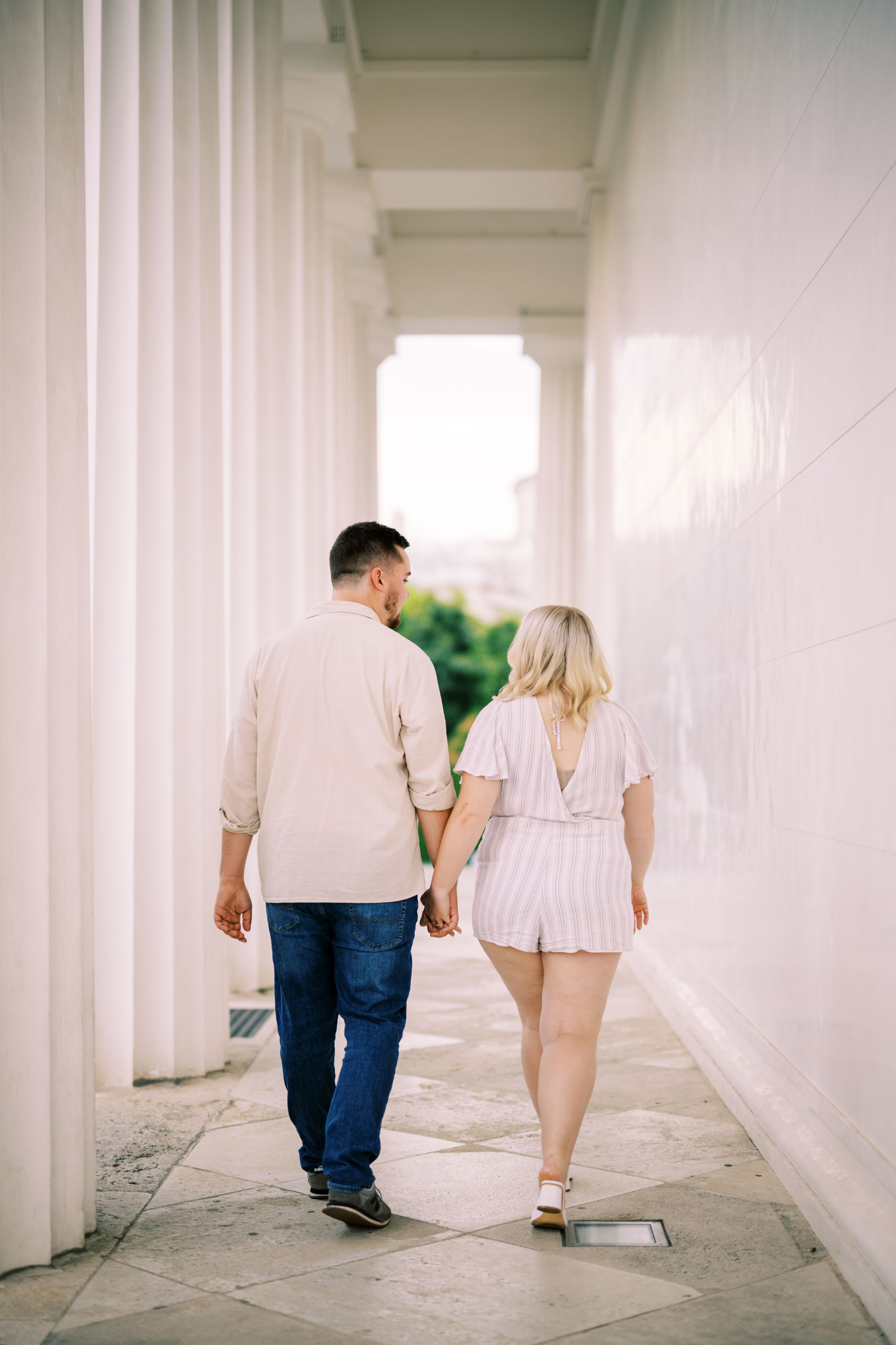 A couple holds hands while walking down a bright white colonnade hallway with stone pillars and sunlit greenery outside.