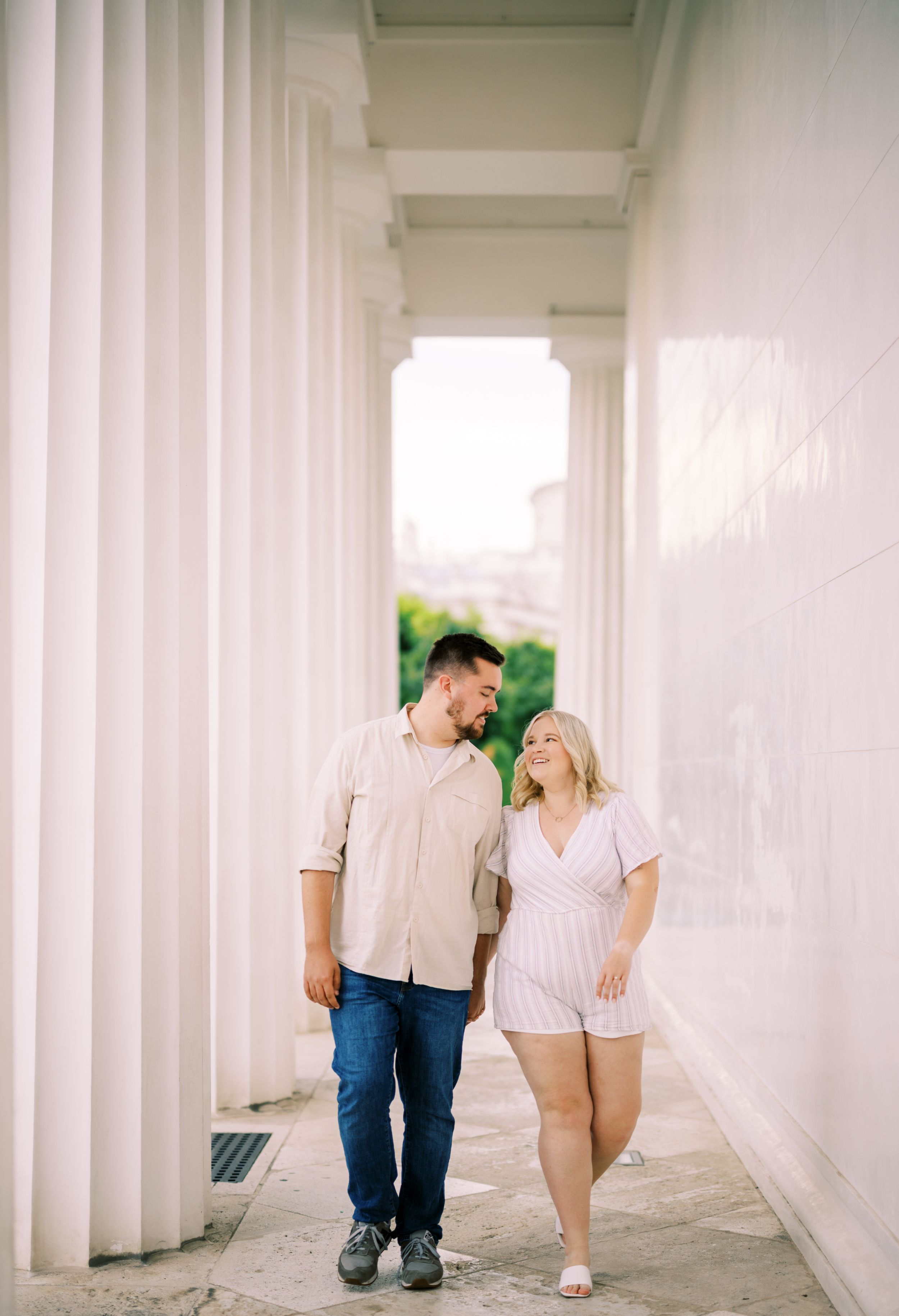 A couple shares a candid moment while walking together through a bright white colonnade hallway.