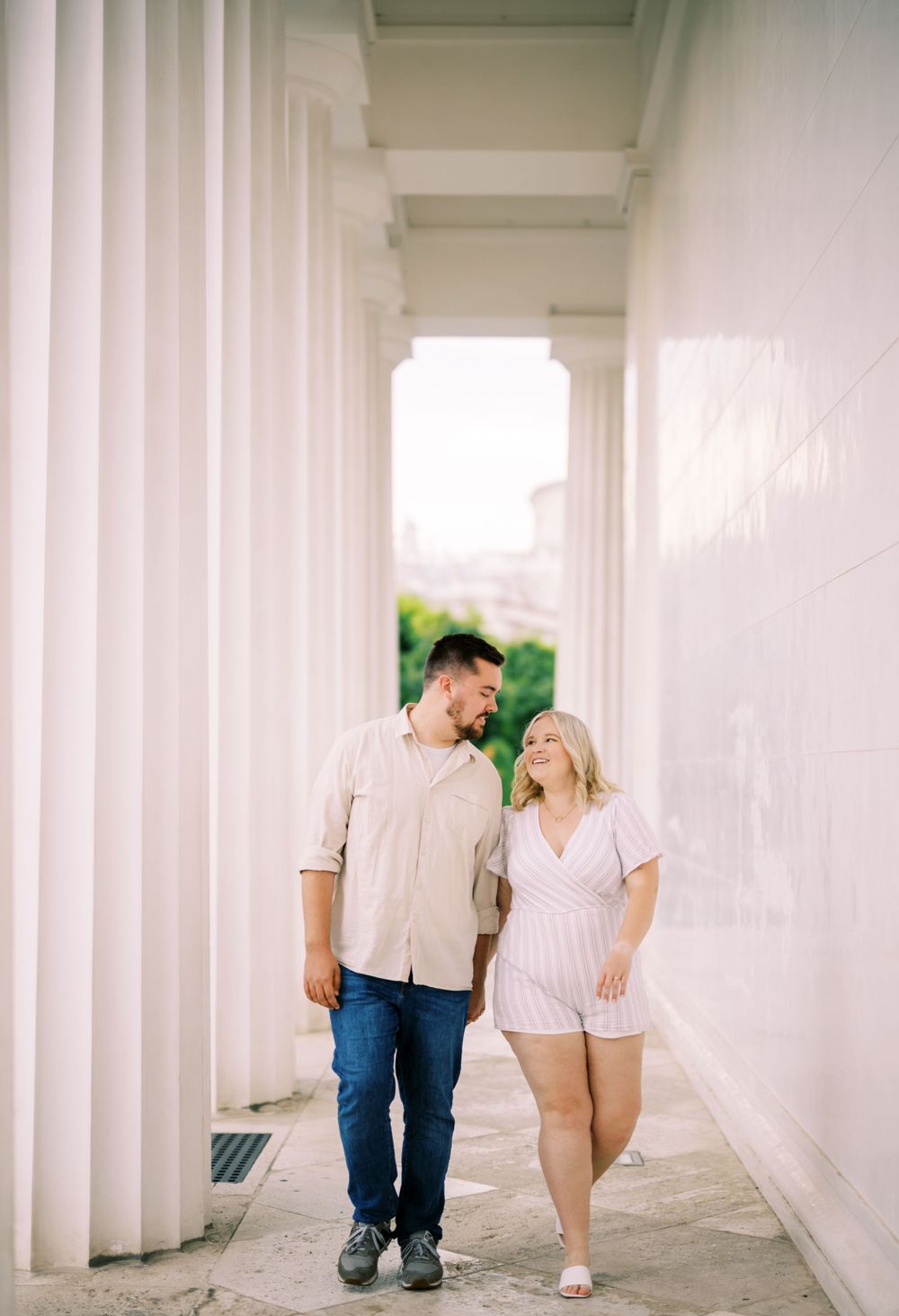 A couple shares a candid moment while walking together through a bright white colonnade hallway.