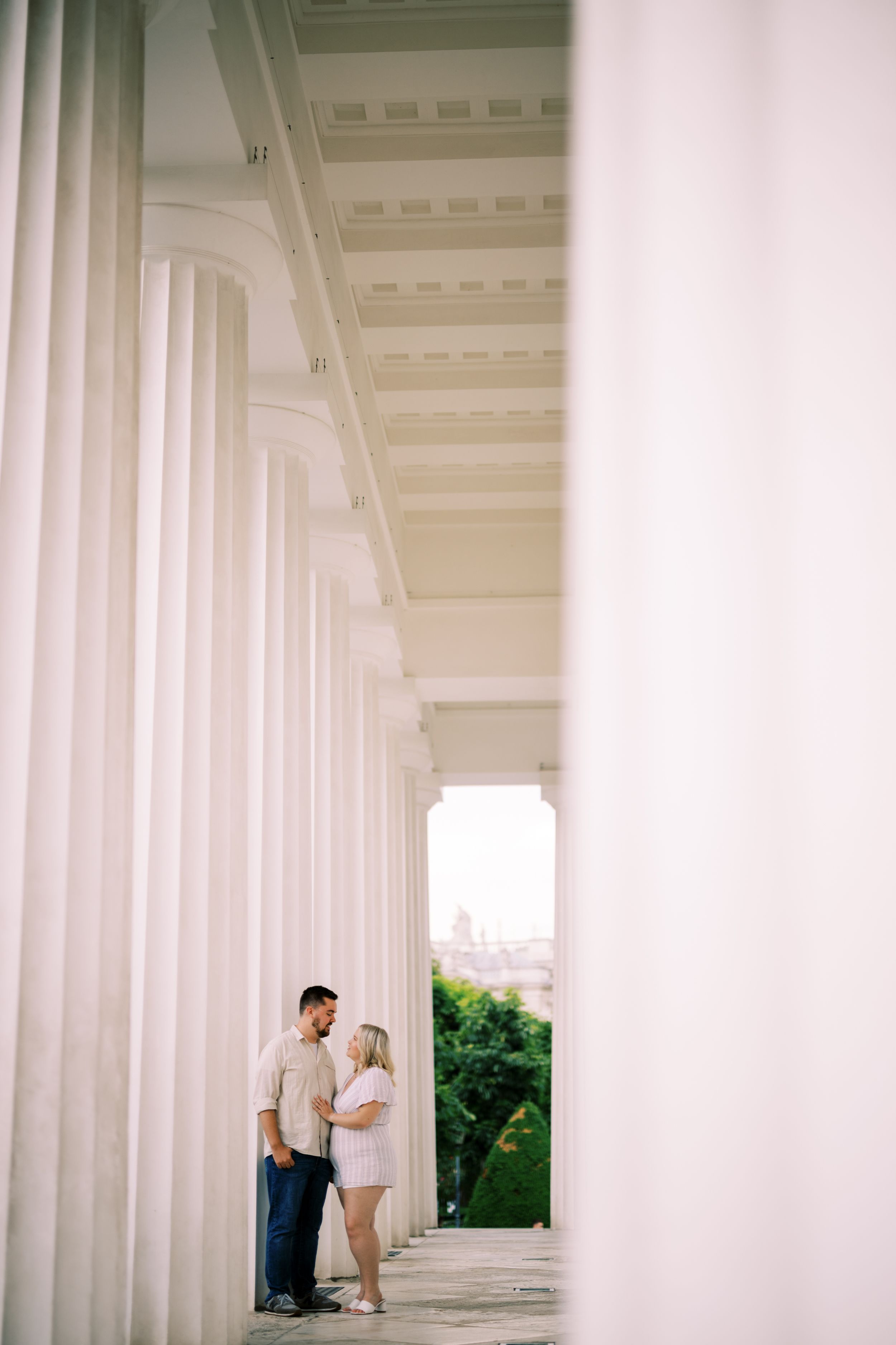 A couple stands intimately between grand white columns of a classical building with greenery visible in the background.