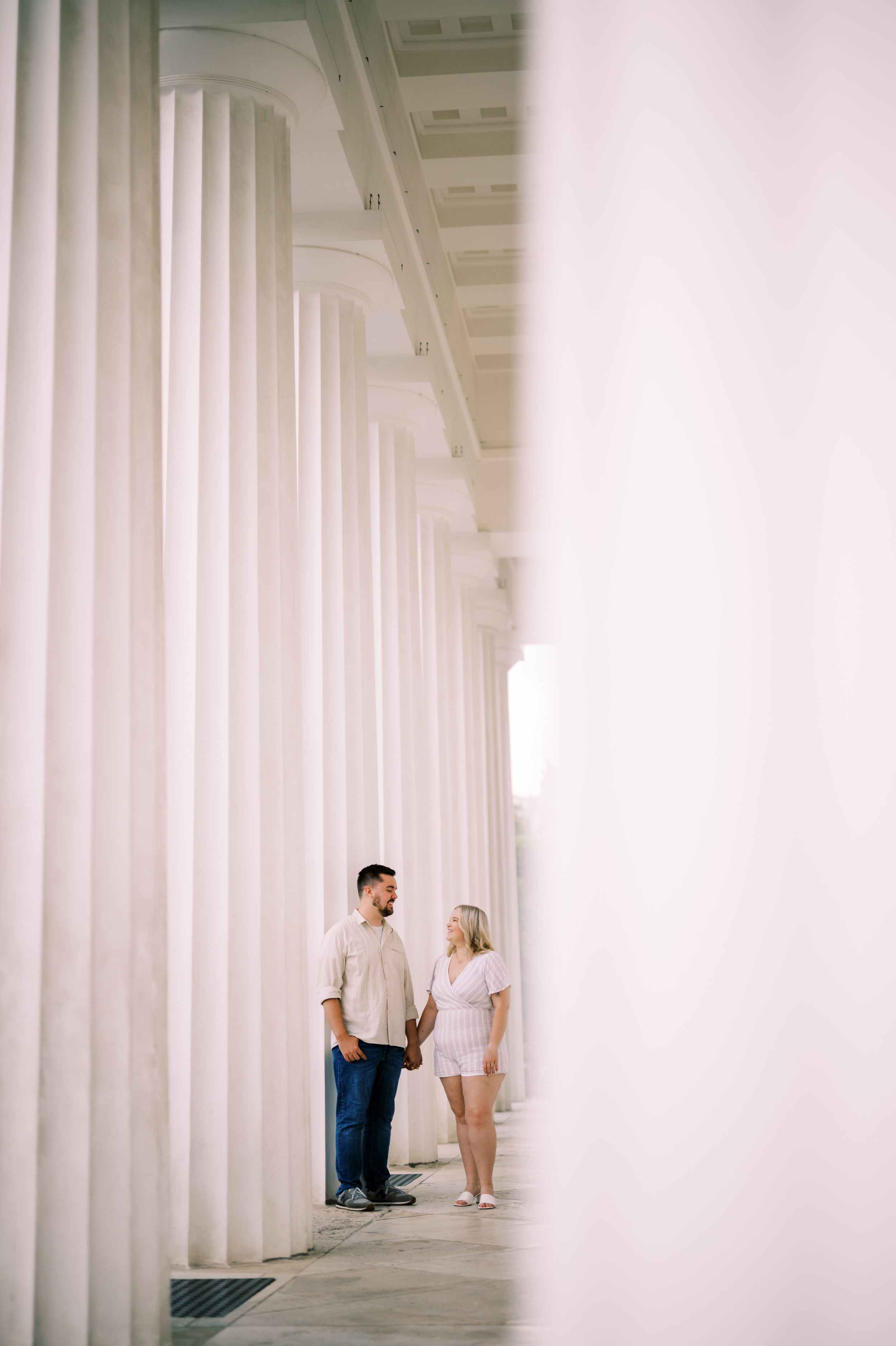 A couple stands together holding hands between tall white columns in an elegant architectural setting.