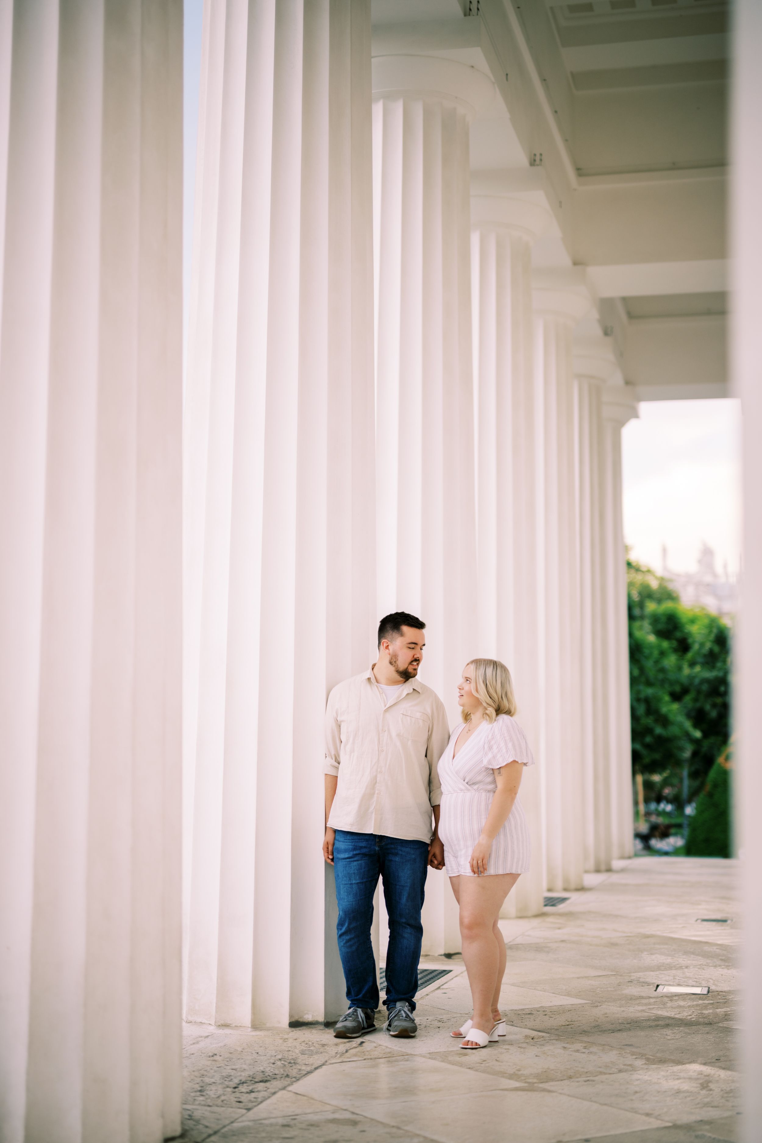 A couple stands between elegant white columns during a romantic photoshoot in a classical architectural setting.