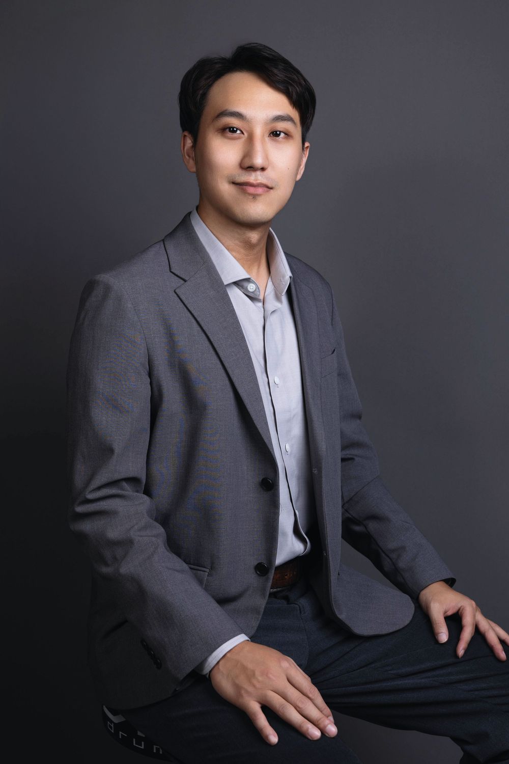 Asian professional man in gray suit jacket seated, confident business portrait against dark gray backdrop.
