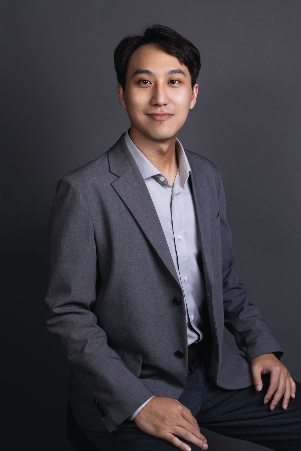 Smiling Asian businessman in gray blazer seated, professional headshot photographed against dark gray studio background.