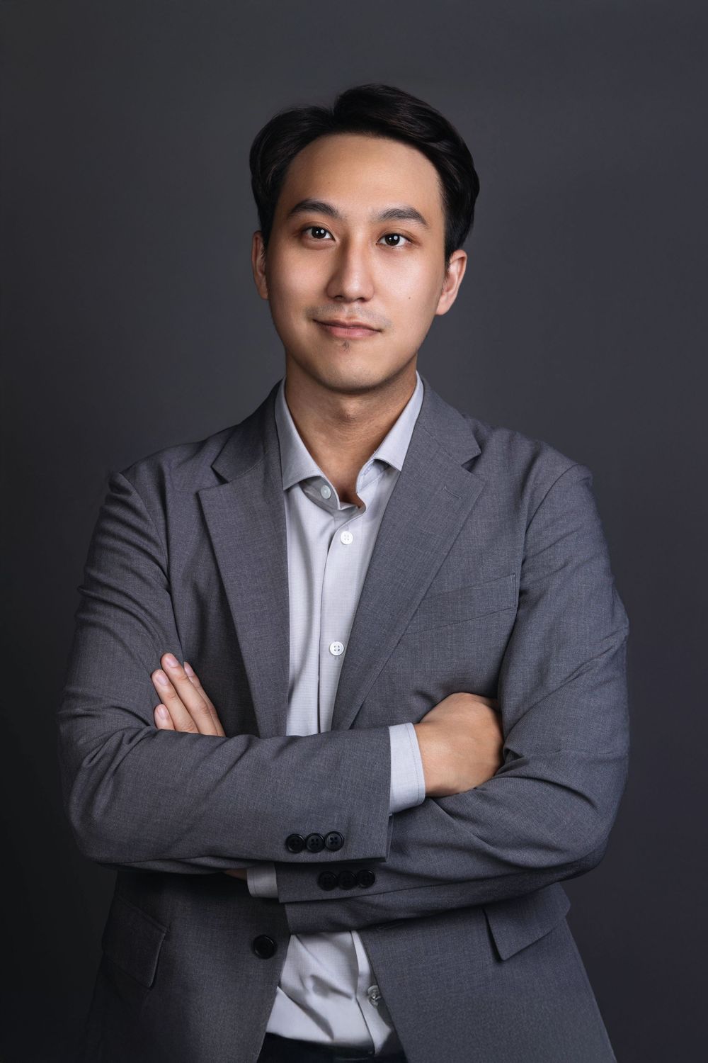 Asian man in gray blazer with arms crossed, confident professional business portrait on dark gray background.