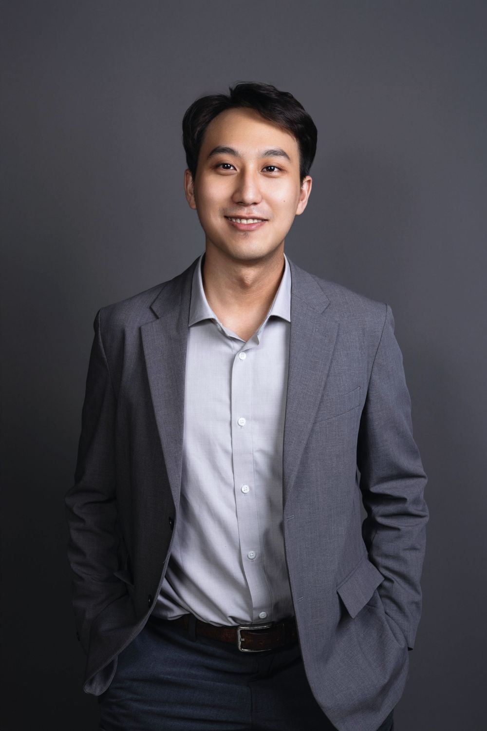 Smiling Asian man in gray suit jacket standing with hands in pockets, professional business portrait.